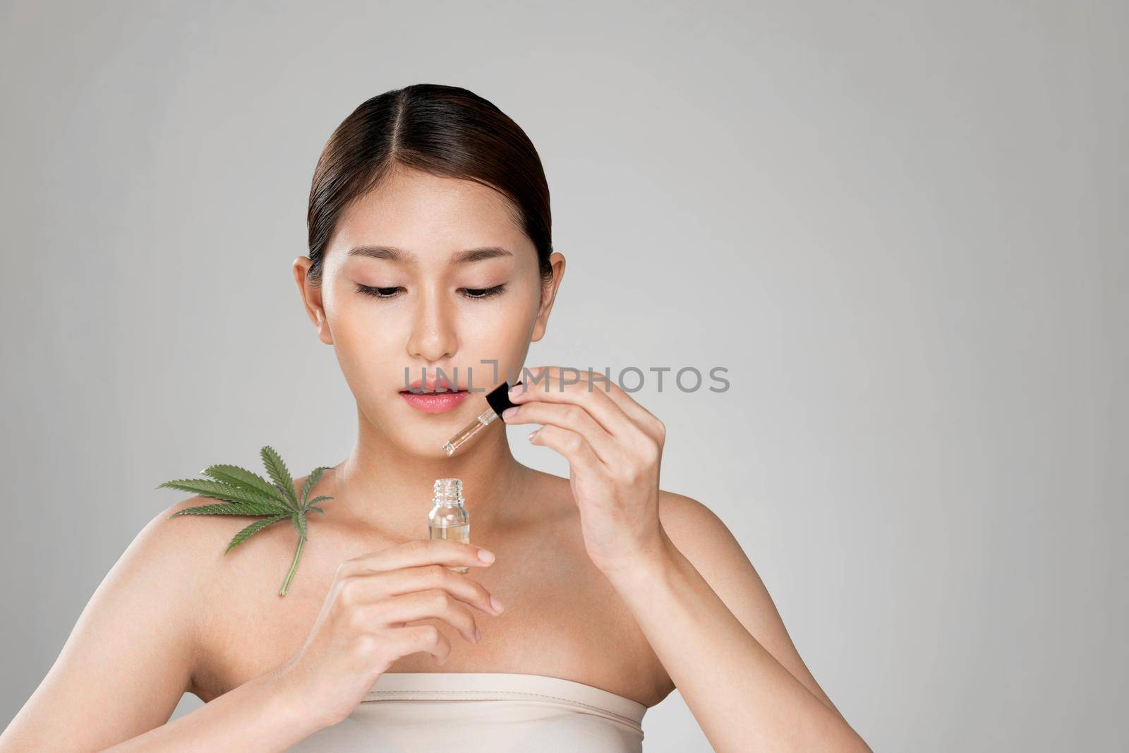 Portrait of young ardent woman with healthy fresh skin holding green hemp leaf and cbd oil. Combination of beauty and cannabis concept.