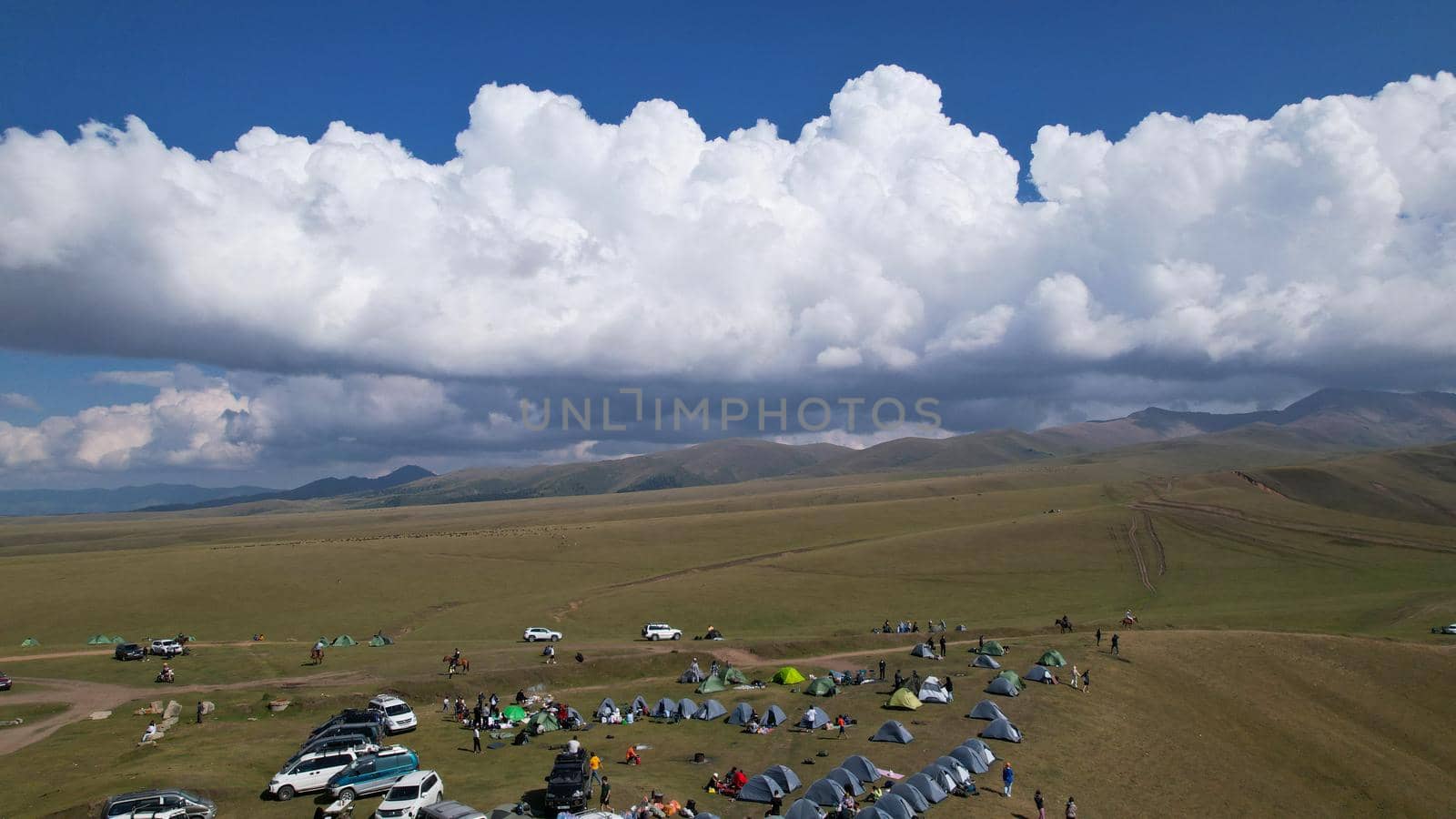 Big white clouds over green hills and mountains. Top view from the drone on endless fields. Roads are visible in places, herds of animals graze. A tent camp has been set up. Coniferous trees in gorge