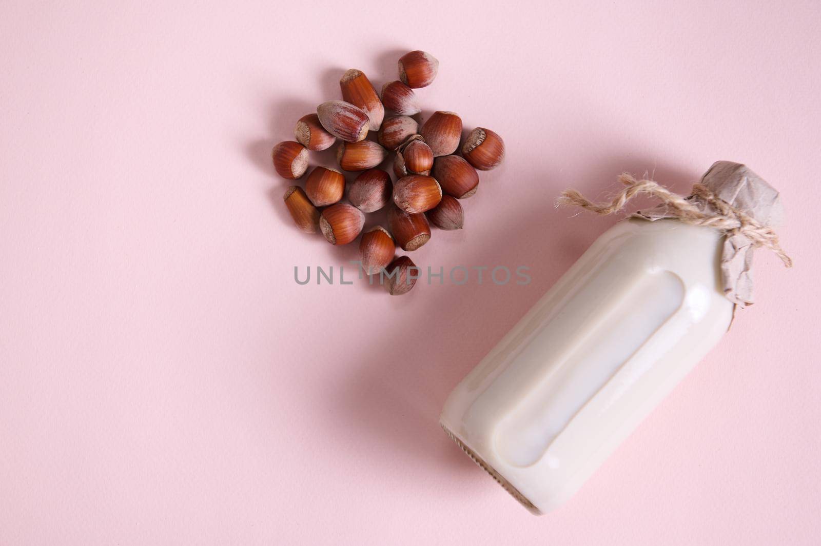 Still life with a plant based milk in a glass bottle, alternative non dairy drink and a heap of organic wholesome hazelnuts on a pink background with advertising copy space. Healthy food and drink