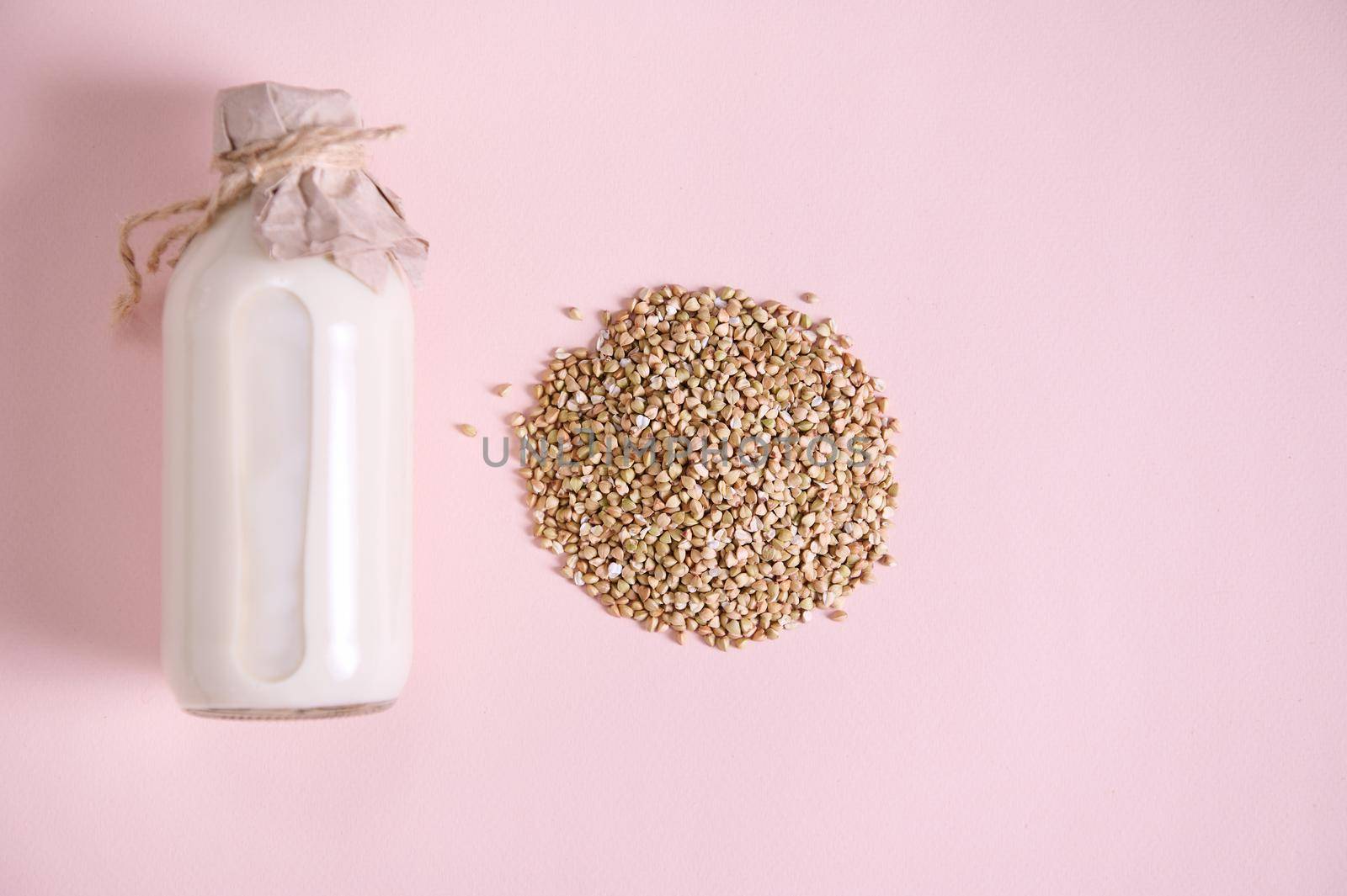 Flat lay. Selective focus. Heap of green buckwheat grains and a bottle of organic wholesome vegan milk, a plant based, non dairy, lactose free drink, milk replacer on pink background. Copy ad space