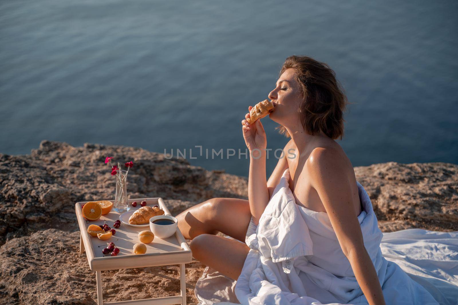 Woman covered with a blanket of bed relaxing and watching the seascape at sunrise. She holds a cup of coffee in her hand in front of her is a table with fruits and croissants. Wanderlust and freedom concept scene