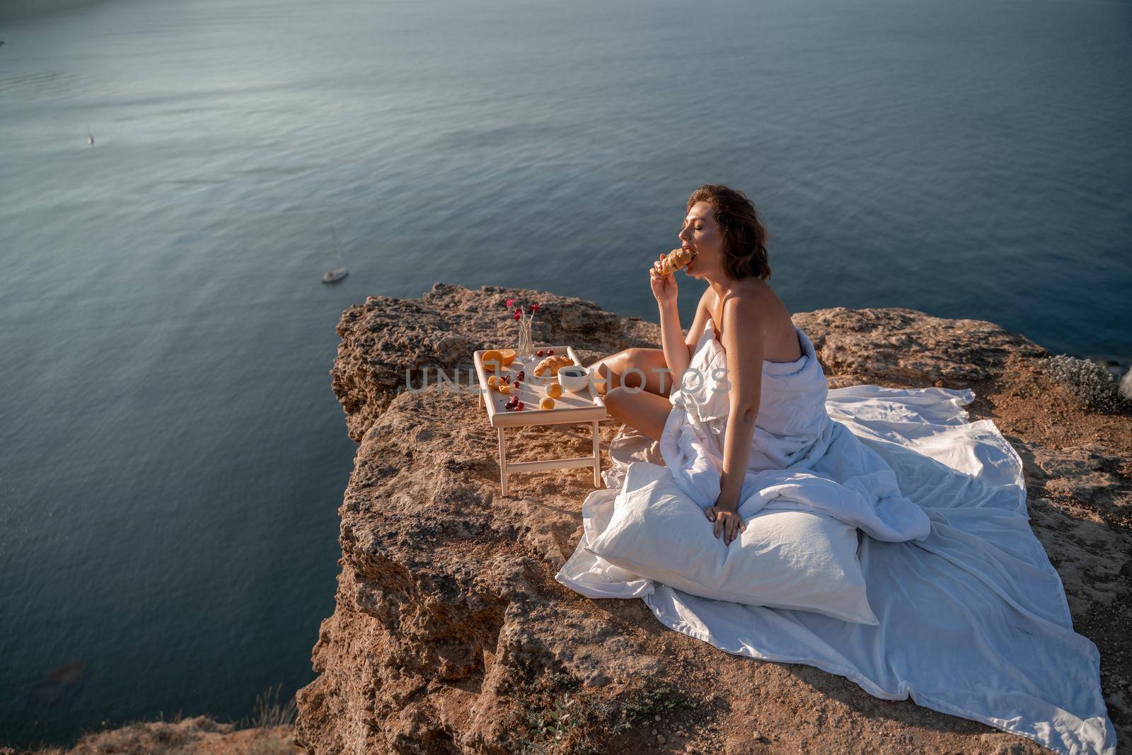 Woman covered with a blanket of bed relaxing and watching the seascape at sunrise. She holds a cup of coffee in her hand in front of her is a table with fruits and croissants. Wanderlust and freedom concept scene