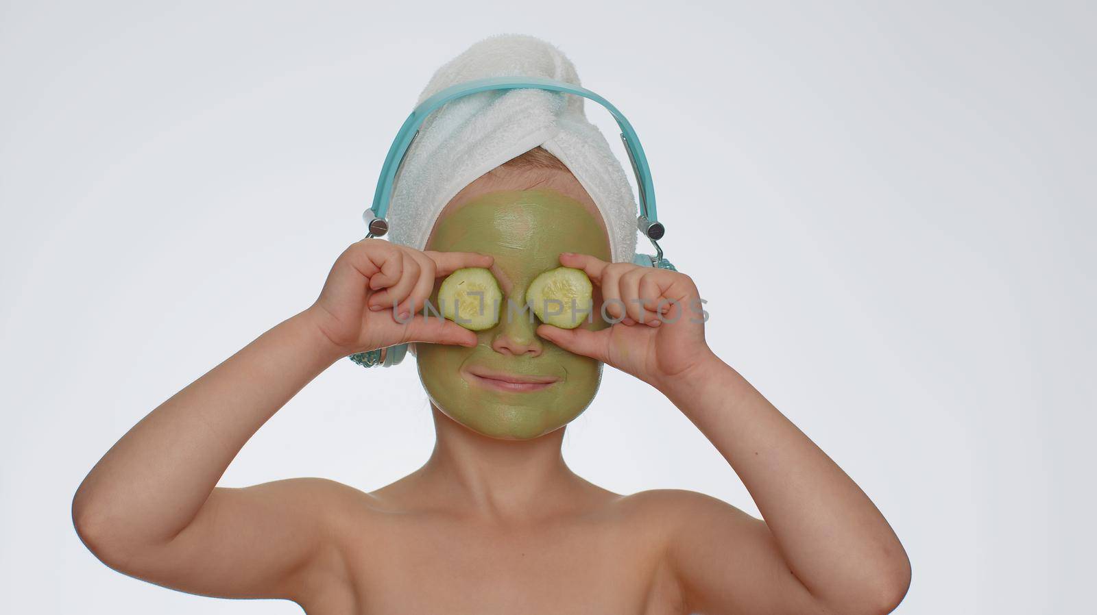 Smiling young child girl after bath in towel on head and moisturizing green cucumber facial mask listening to music on headphones, dancing. Female teenager kid skin care treatment, natural cosmetics