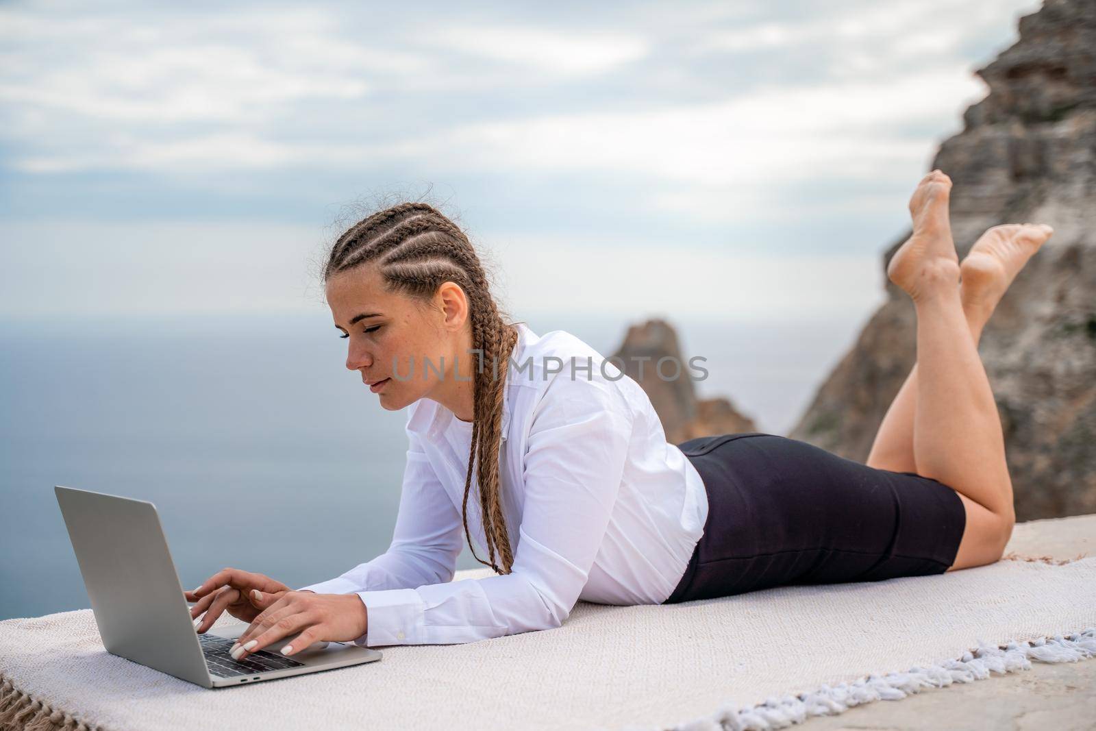 A woman is lying and typing on a laptop keyboard on a terrace with a beautiful sea view. Wearing a white blouse and black skirt. Freelance travel and vacation concept, digital nomad. by Matiunina