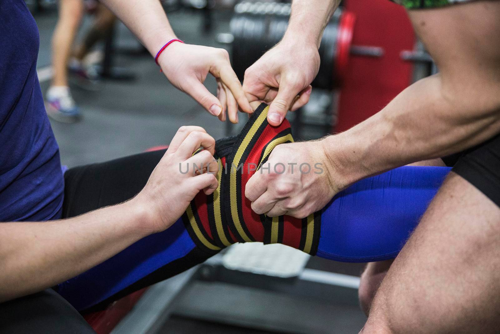 Sportsman is bandages his partner's knee by elastic bandage in the gym. Squat with a barbell.