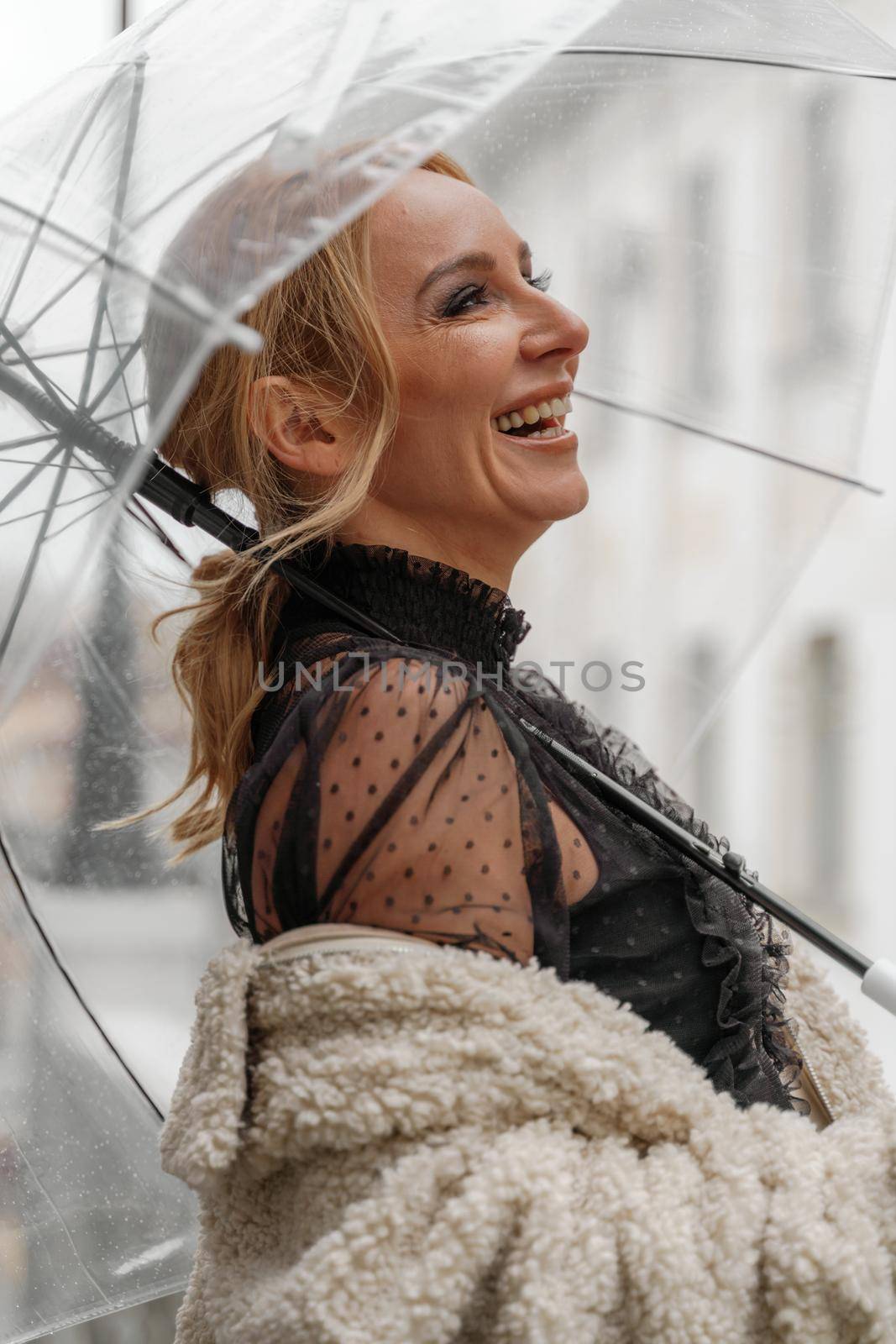 The blonde stands under a transparent umbrella during the rain. The fall season. Rear view. The woman is dressed in a black lace dress, her hair pulled back in a ponytail