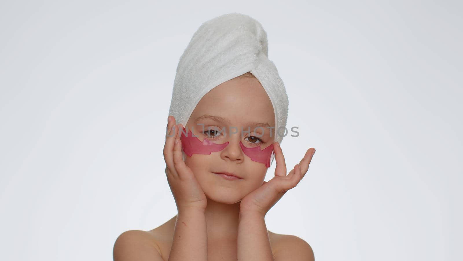 Beautiful young smiling child girl in towel on head applying pink patches under eyes. Teenager kid face skin care treatment, natural cosmetics. Female portrait on white background. Perfect fresh clean