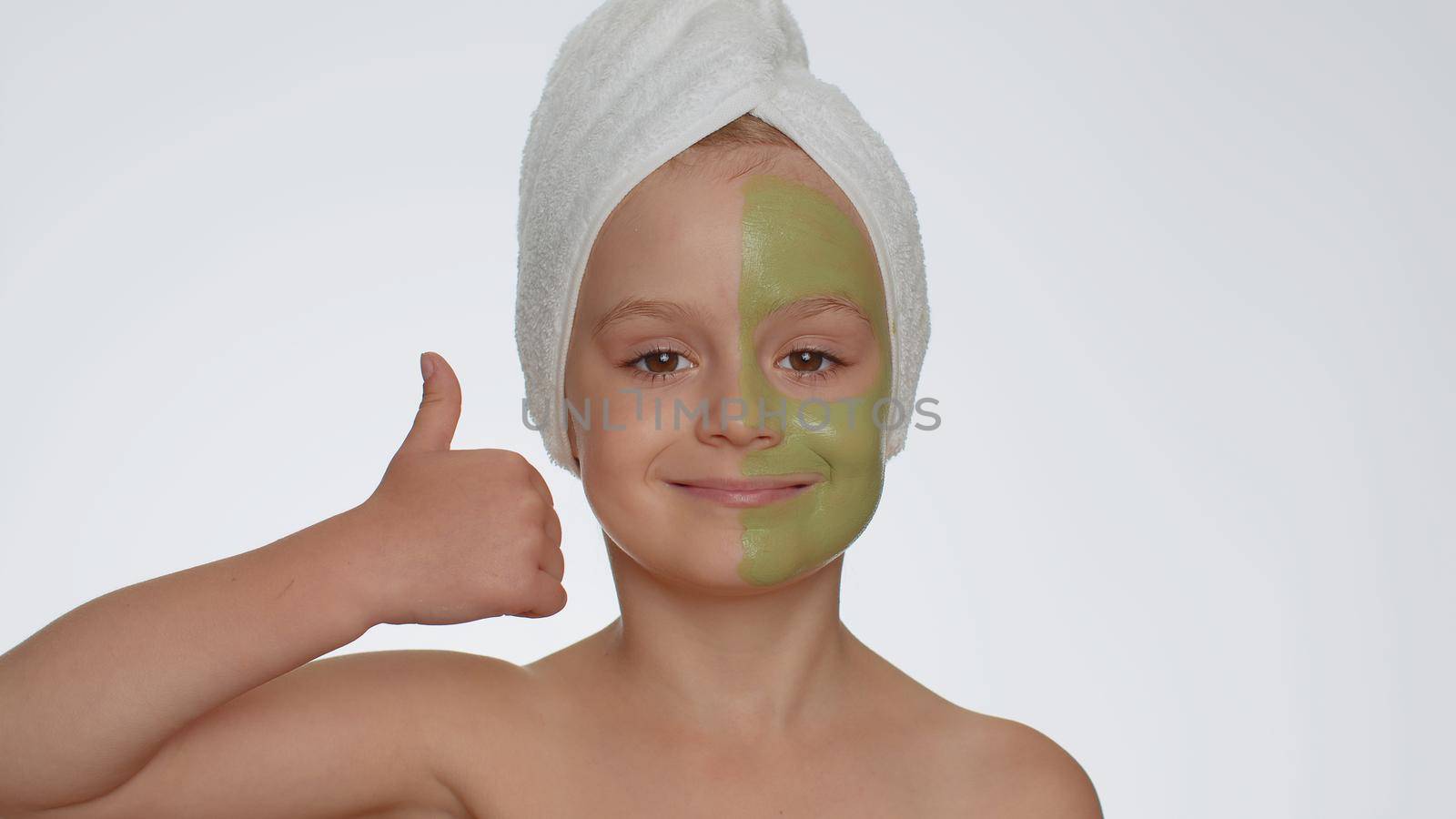 Lovely pretty young smiling child girl applying cleansing moisturizing green vitamin cucumber facial mask, showing thumbs up. Teenager kid face skin care treatment, natural cosmetics. Female portrait