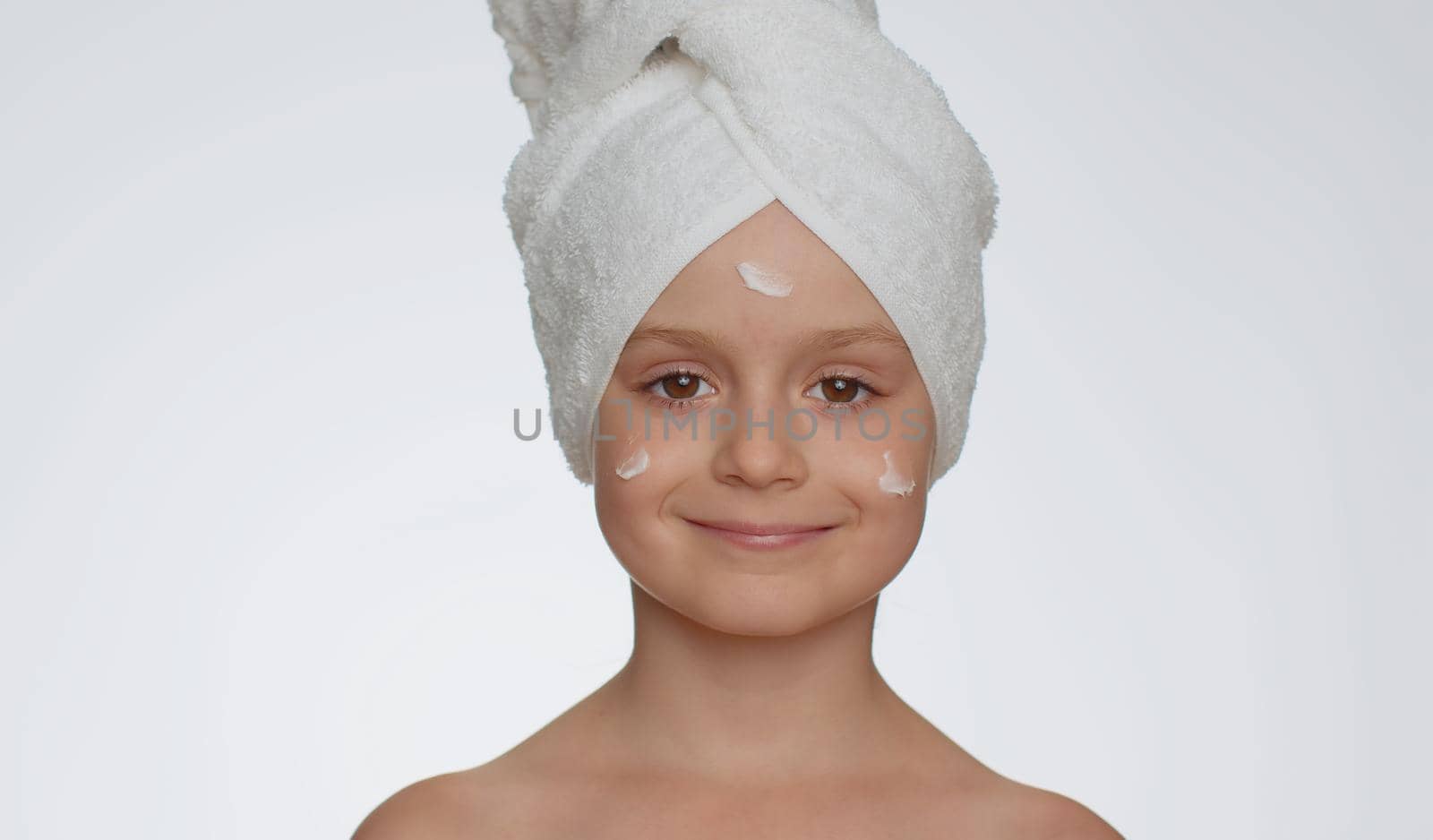 Lovely young child girl after bath in towel on head applying cleansing moisturizing cream. Teenager kid face skincare healthy treatment, natural creme cosmetics. Female portrait. Perfect fresh clean