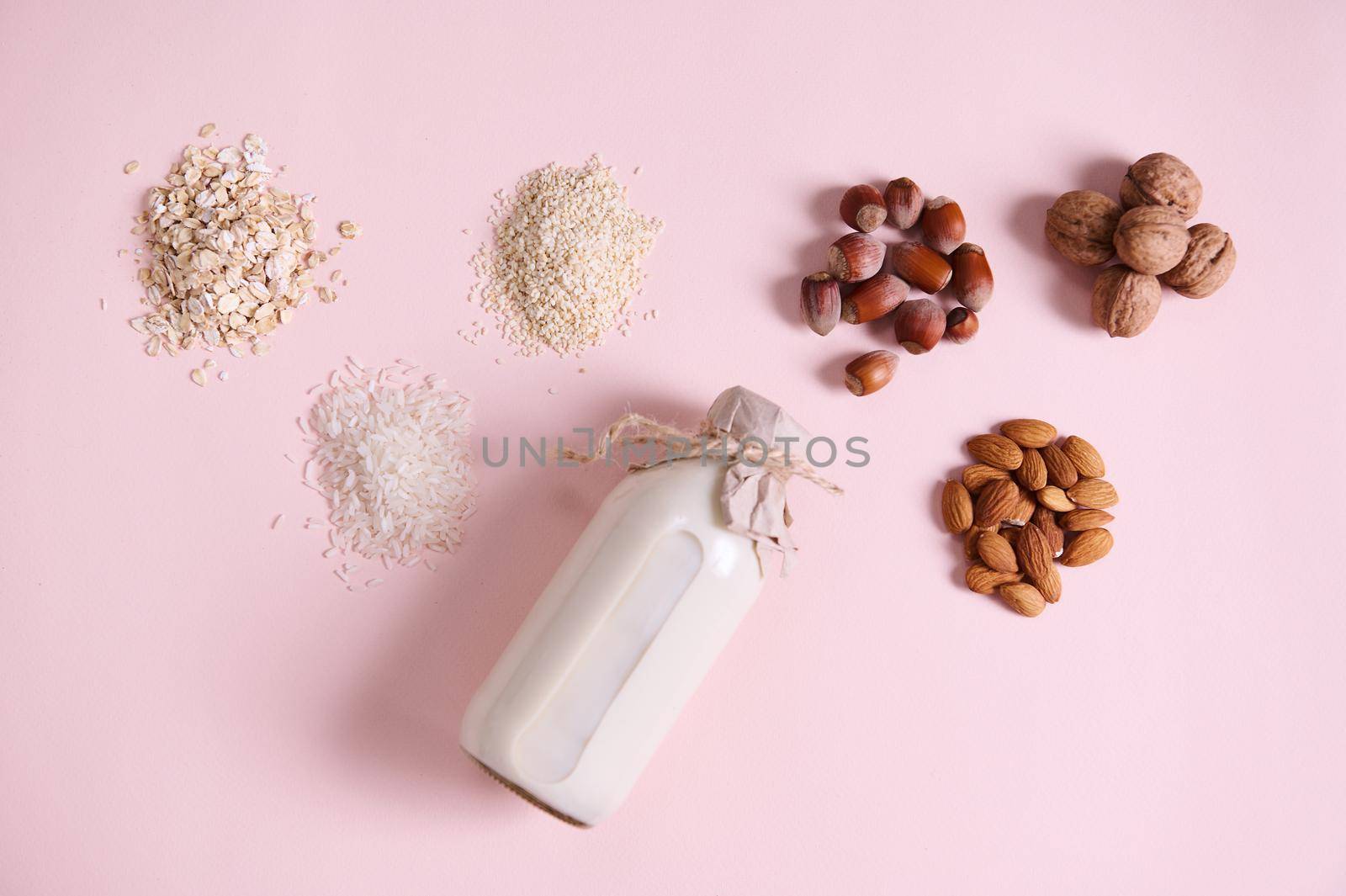 Flat lay. Still life with an assortment of wholesome organic seeds, nut, beans: almond, hazelnut, walnut, sesame, rice, oatmeal and a bottle with vegan plant based milk, isolated on pink background