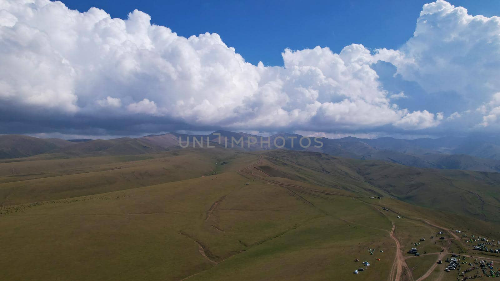 Big white clouds over green hills and mountains. Top view from the drone on endless fields. Roads are visible in places, herds of animals graze. A tent camp has been set up. Coniferous trees in gorge