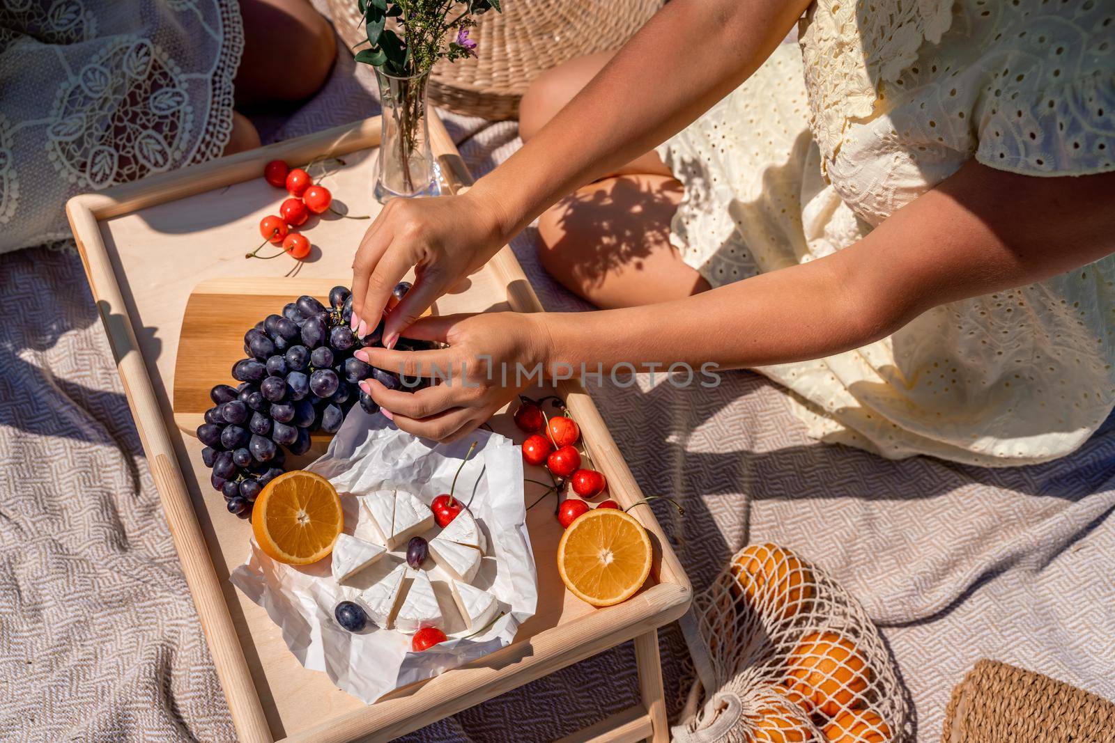 Romantic picnic for two with fruit, bread and cheese. Oranges, cherries, black grapes and camembert on a wooden table. The girl's hands with a manicure tear off a grape berry sitting on a light blanket
