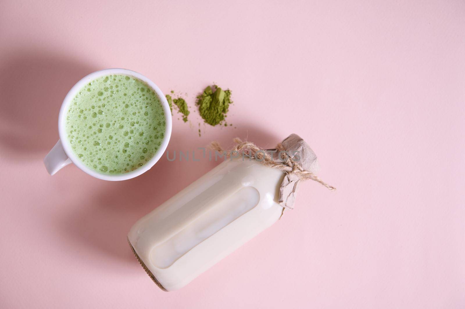 Still life. Bottle of organic plant based milk on pink surface near white ceramic cup of antioxidant healthy drink- whipped matcha latte with foam and a heap of powdered japanese green tea. Copy space