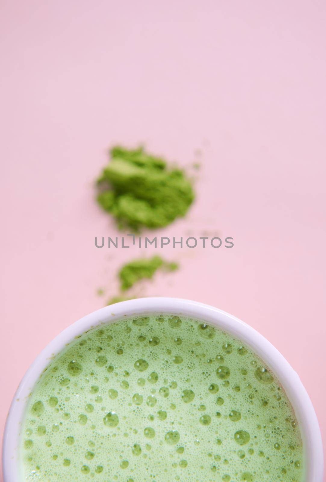 Selective focus on a white cup of whipped green tea with foam and blurred powdered japanese matcha, over pink background with copy advertising space. Antioxidant healthy drink. Oriental traditions