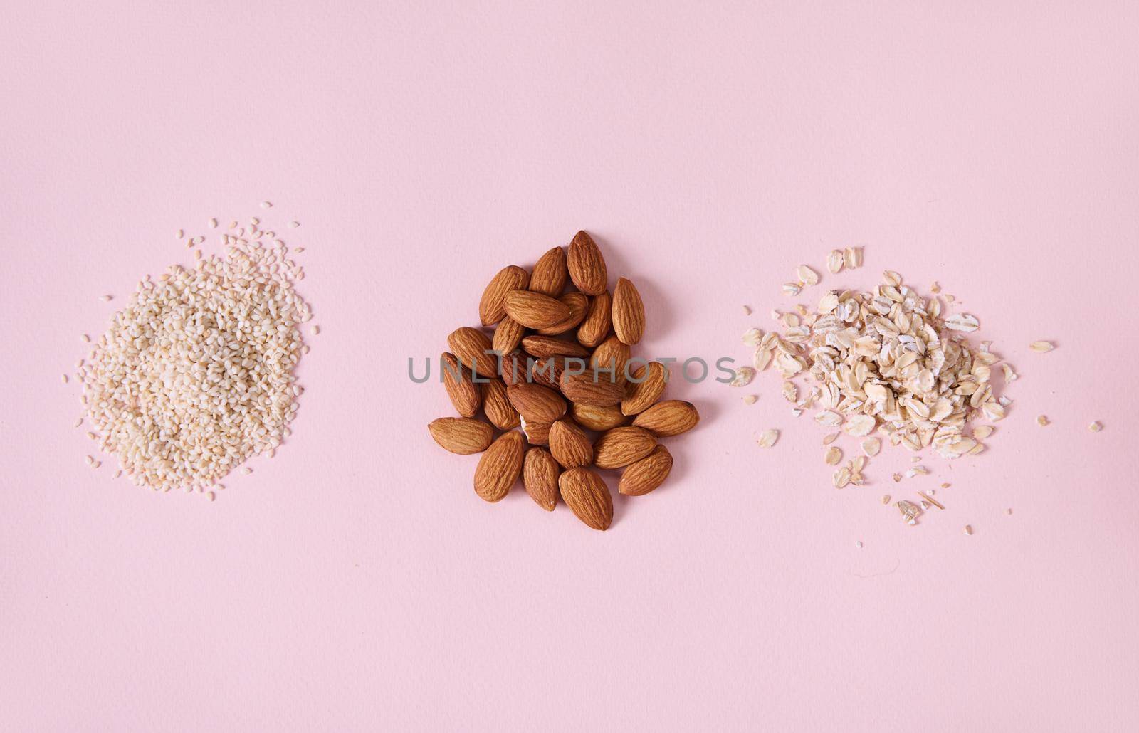 Horizontal studio shot of healthy raw organic almond nuts, sesame seeds and oat-flakes scattered on pink background by artgf