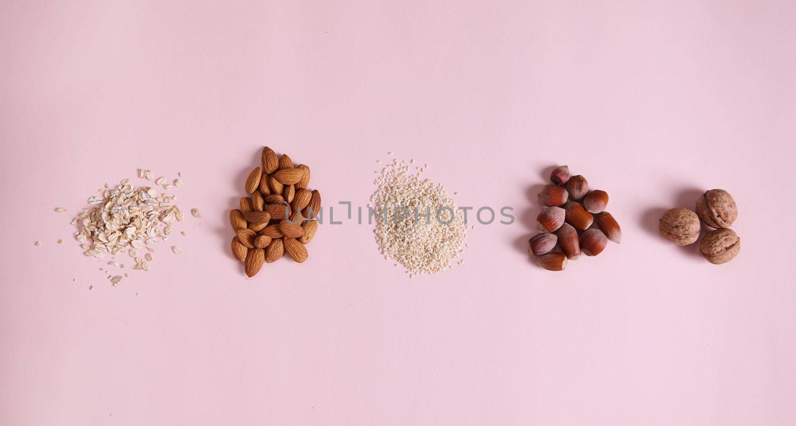 Flat lay. Stacks of organic sesame seeds, almond nuts, oat-flakes, walnut and hazelnut scattered on pink background by artgf