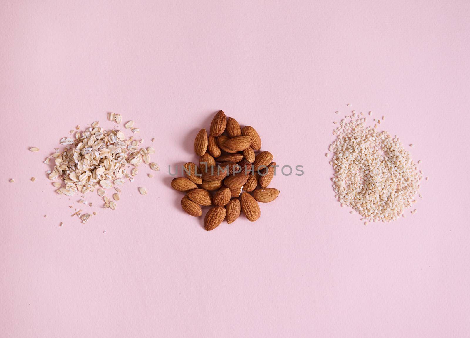 Flat lay of stacks of organic raw vegan superfood: almonds, sesame seeds and oat-flakes scattered on pink background by artgf