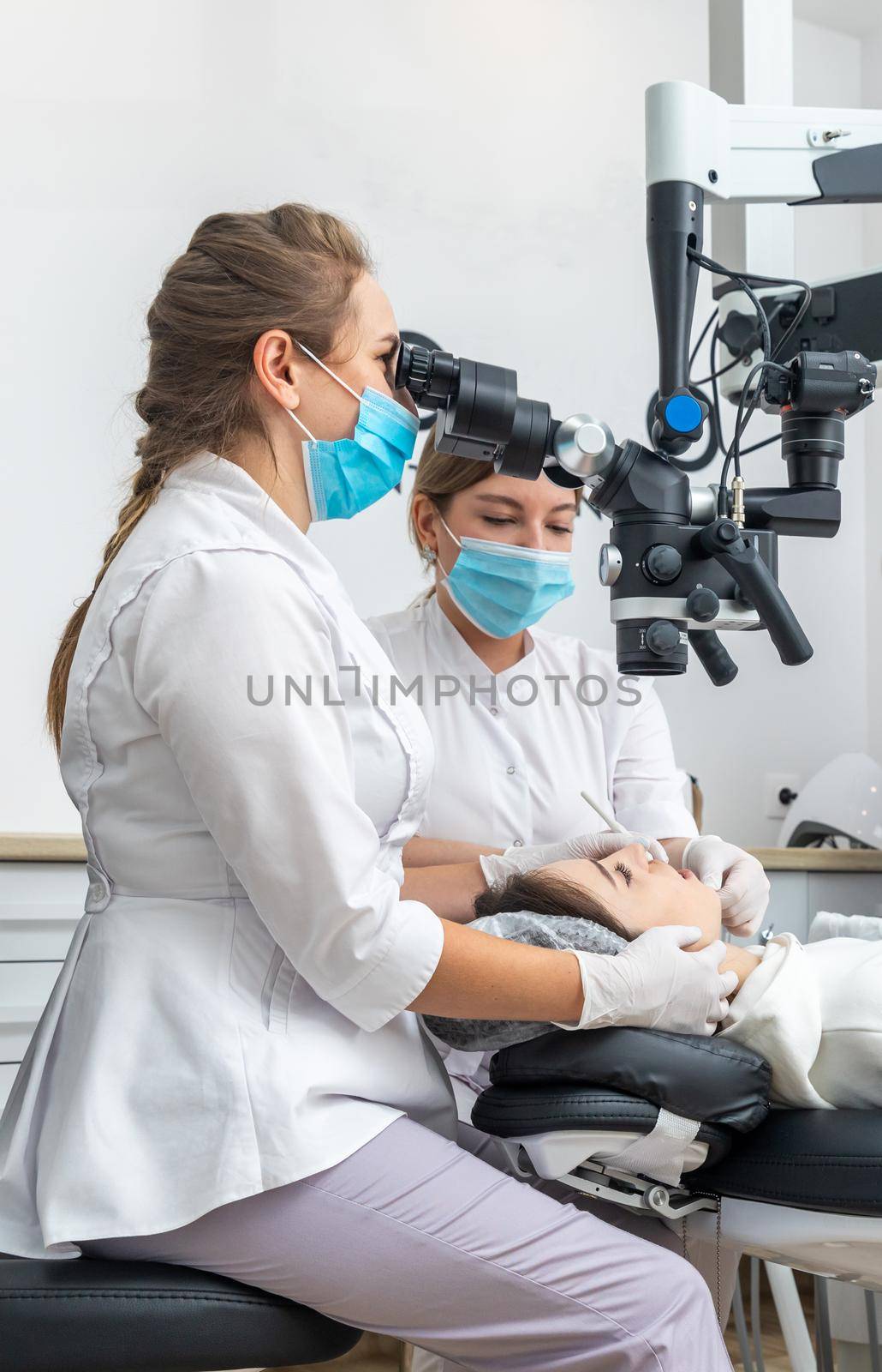 Female dentist using dental microscope treating patient teeth at dental clinic office by Mariakray
