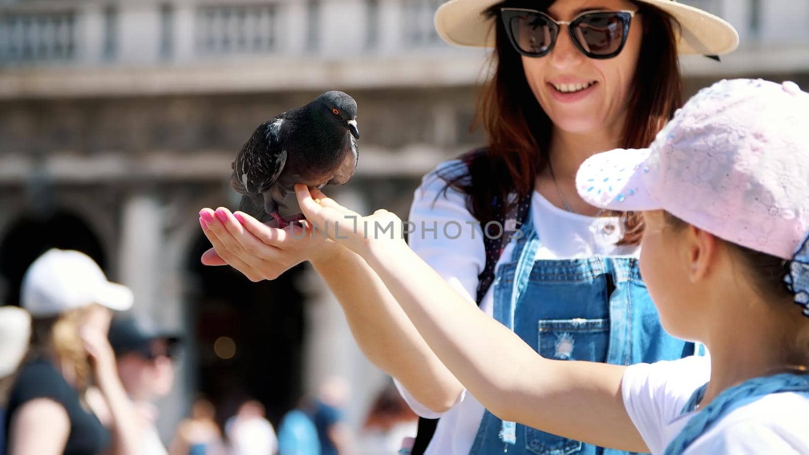 VENICE, ITALY - JULY 7, 2018: view of happy woman and kid girl, tourists, holding pigeons, feeding, play with them, having fun on Piazza San Marco, St Mark's Basilica, on a summer day. High quality photo