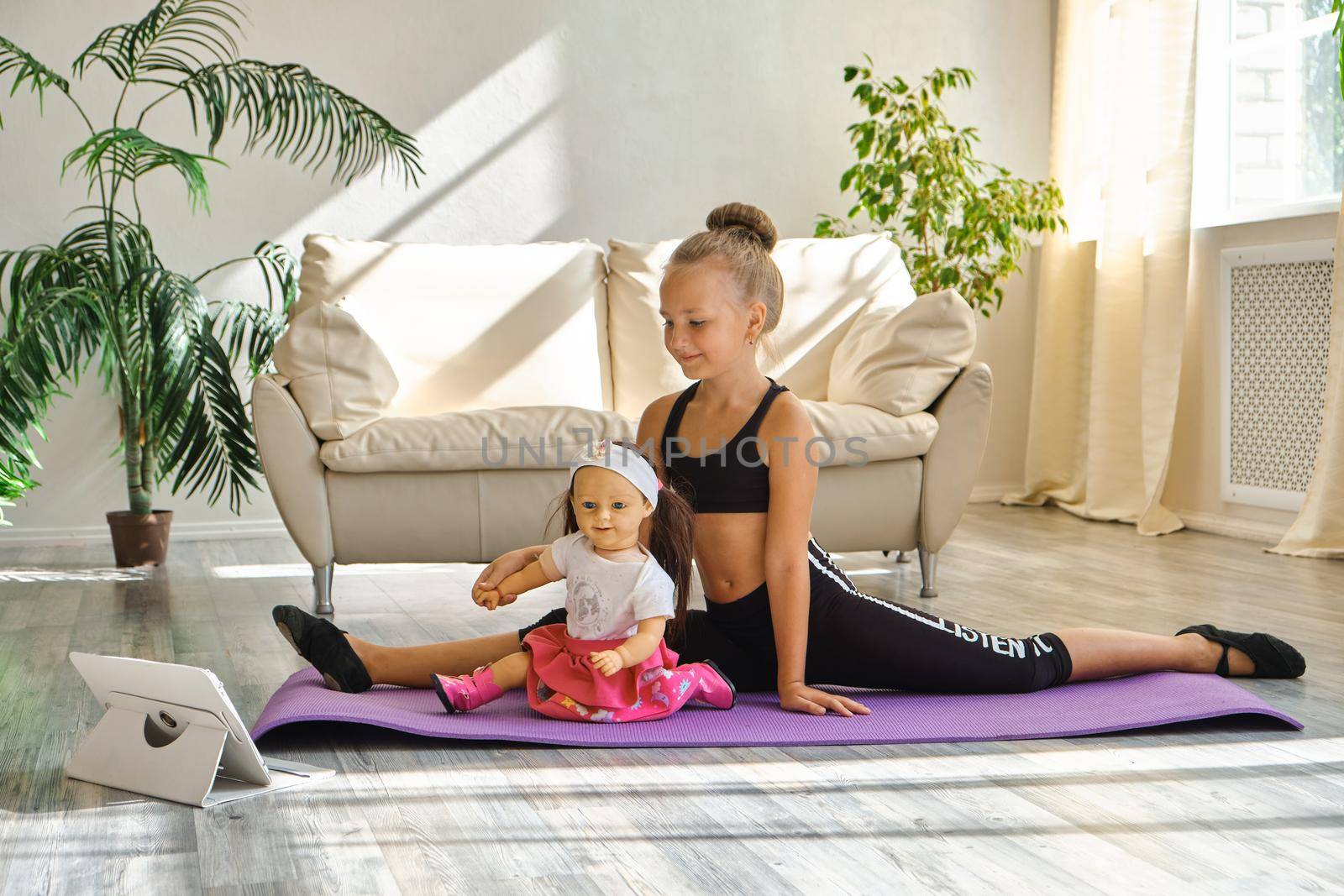 Little cute girl is practicing gymnastics at home. Video lesson. Kid repeating exercises while watching online yoga gymnastics session. Beautiful girl doing fitness exercise at home