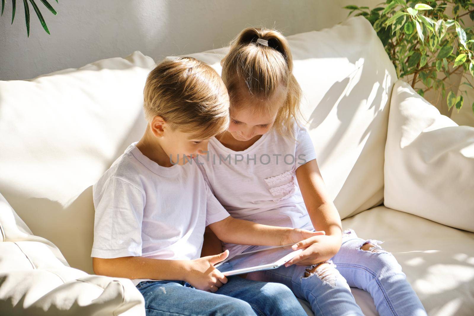 family, children, technology and home concept - smiling brother and sister with tablet pc computer on the sofa in living room