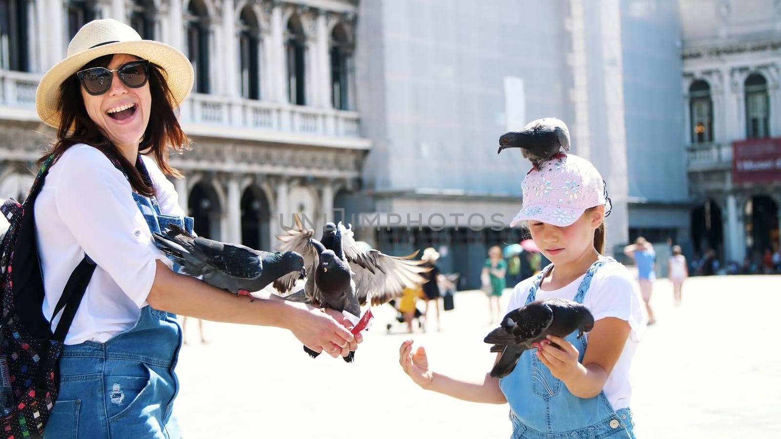 VENICE, ITALY - JULY 7, 2018: view of happy woman and kid girl, tourists, holding pigeons, feeding, play with them, having fun on Piazza San Marco, St Mark's Basilica, on a summer day. High quality photo