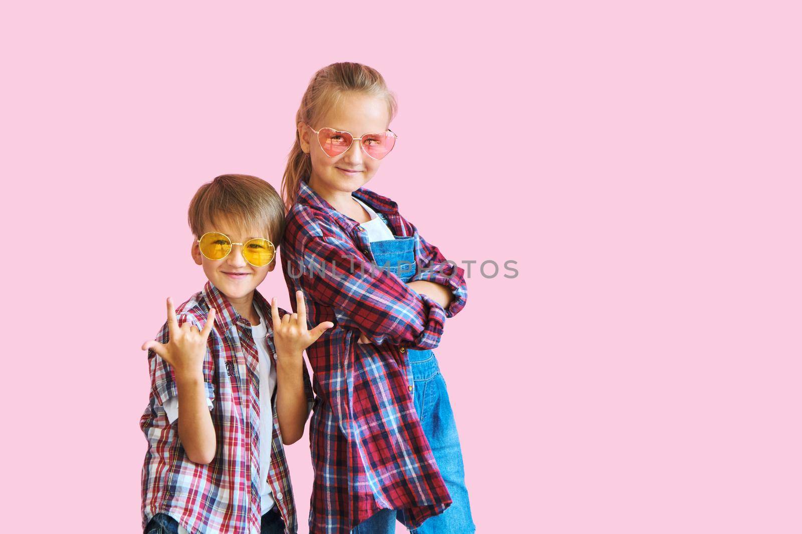 Cute stylish little girl and boy in color sunglasses dressed in plaid shirts, looking at camera and laughing while standing on pink background with copy space.