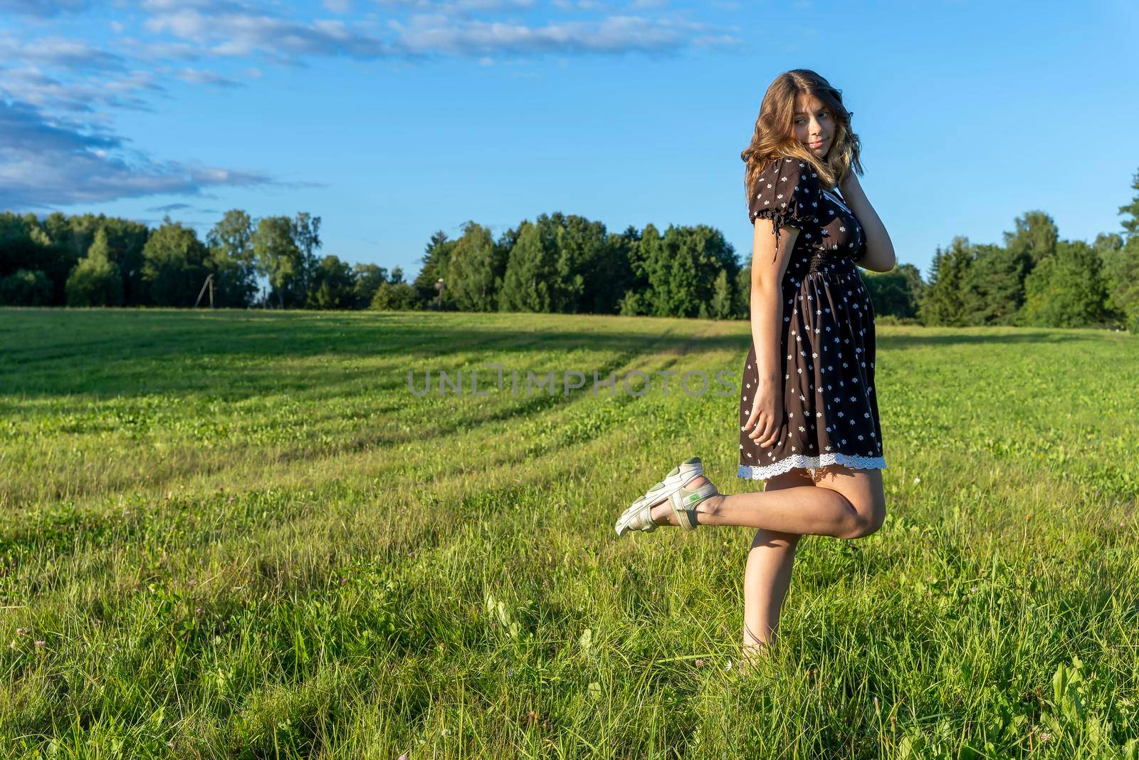 A full-length portrait of an attractive Caucasian girl with long hair in a fashionable short brown dress against a green field in summer in the setting sun. A stylish teenage girl stands on one leg, tucking the other. Attractive young lady looks at the camera