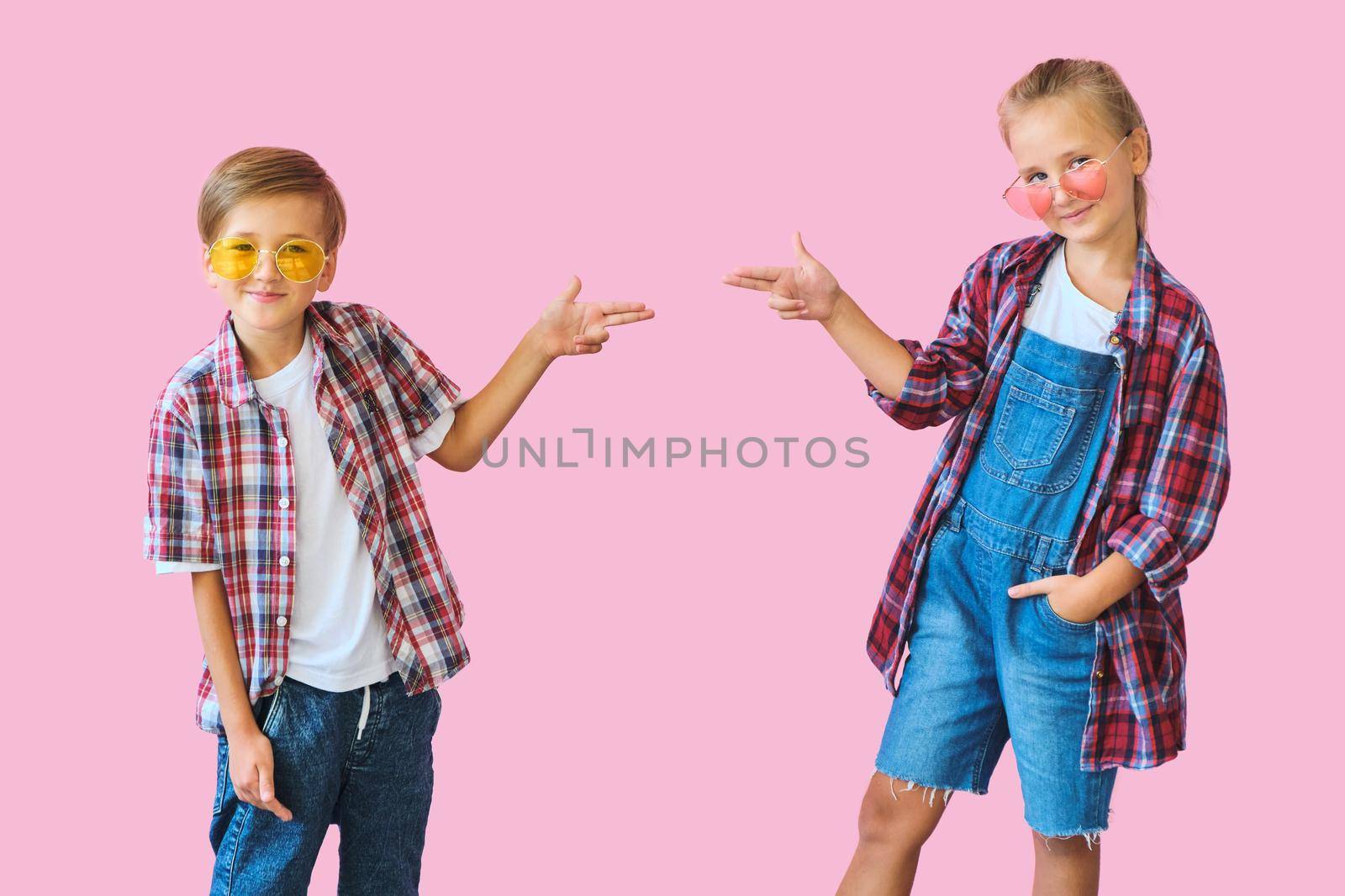 Cute stylish little girl and boy in color sunglasses dressed in plaid shirts, looking at camera and laughing while standing on pink background with copy space.