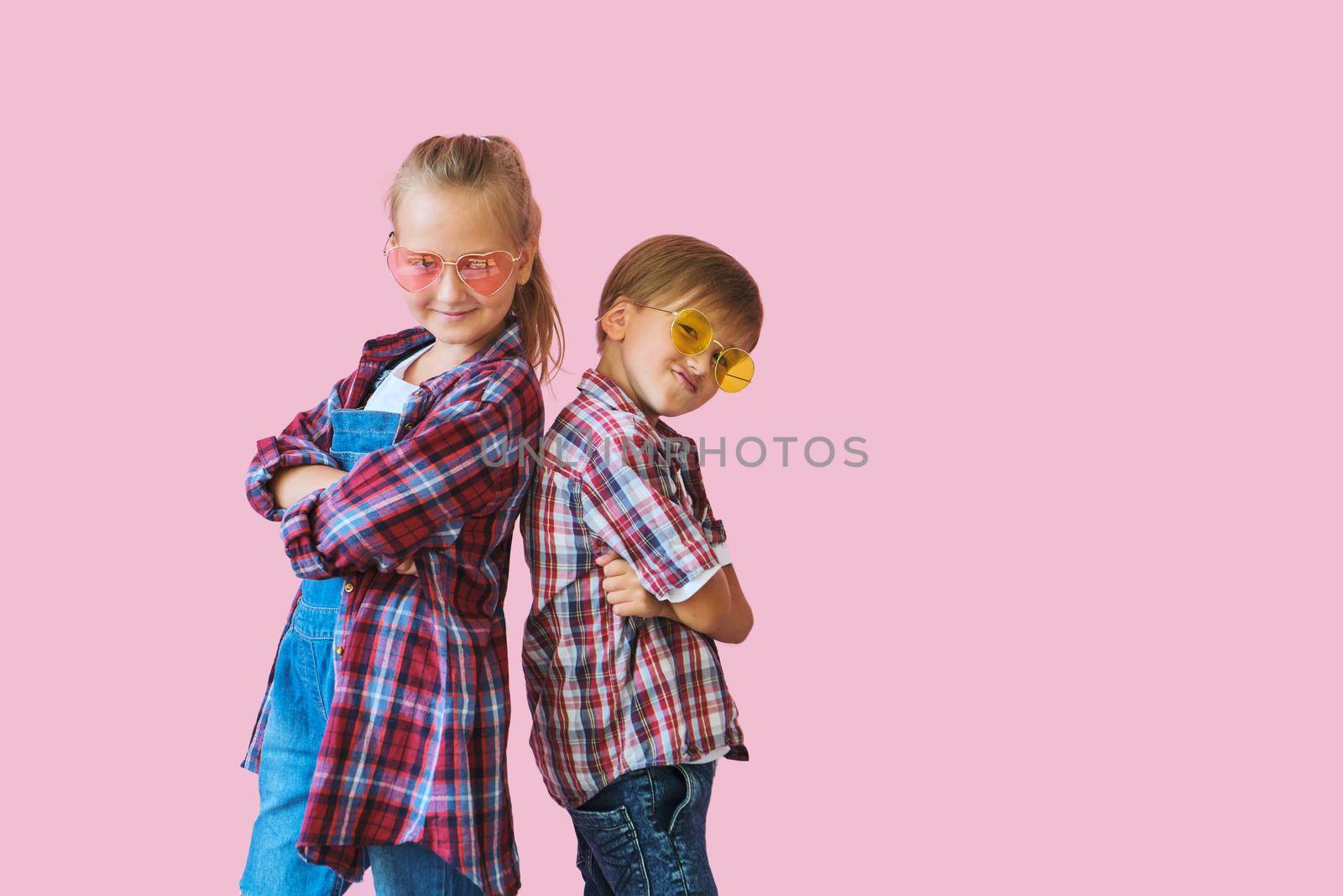 Cute stylish little girl and boy in color sunglasses dressed in plaid shirts, looking at camera and laughing while standing on pink background with copy space.