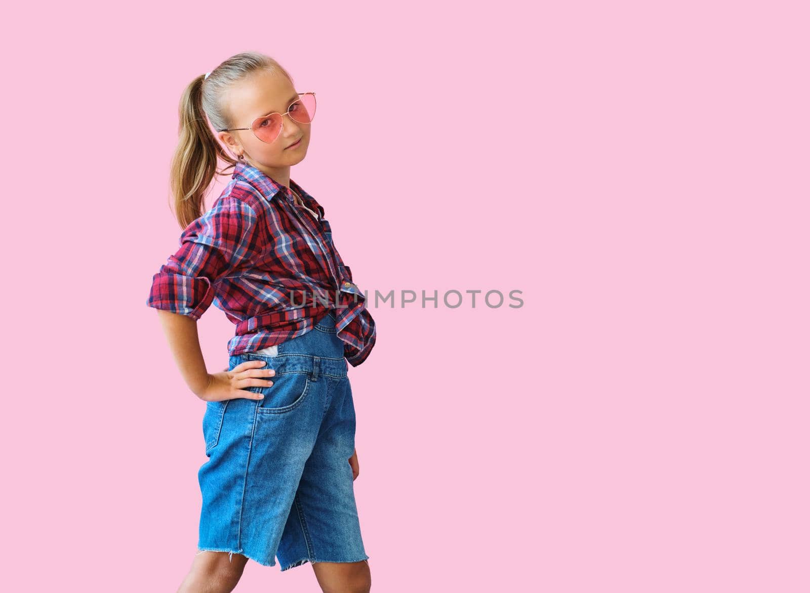 Pretty cool kid girl in pink sunglasses posing against pink background. Fashion style portrait of young happy smiling woman, copy space.