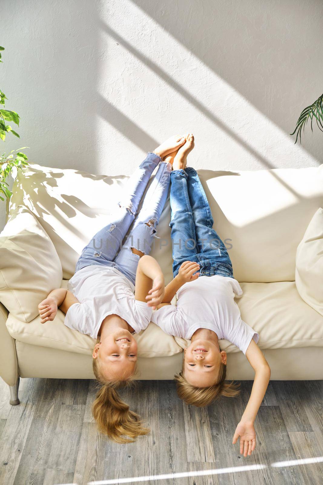 Pretty little girl and boy are lying on their backs on sofa in living room, looking at camera and smiling while playing at home