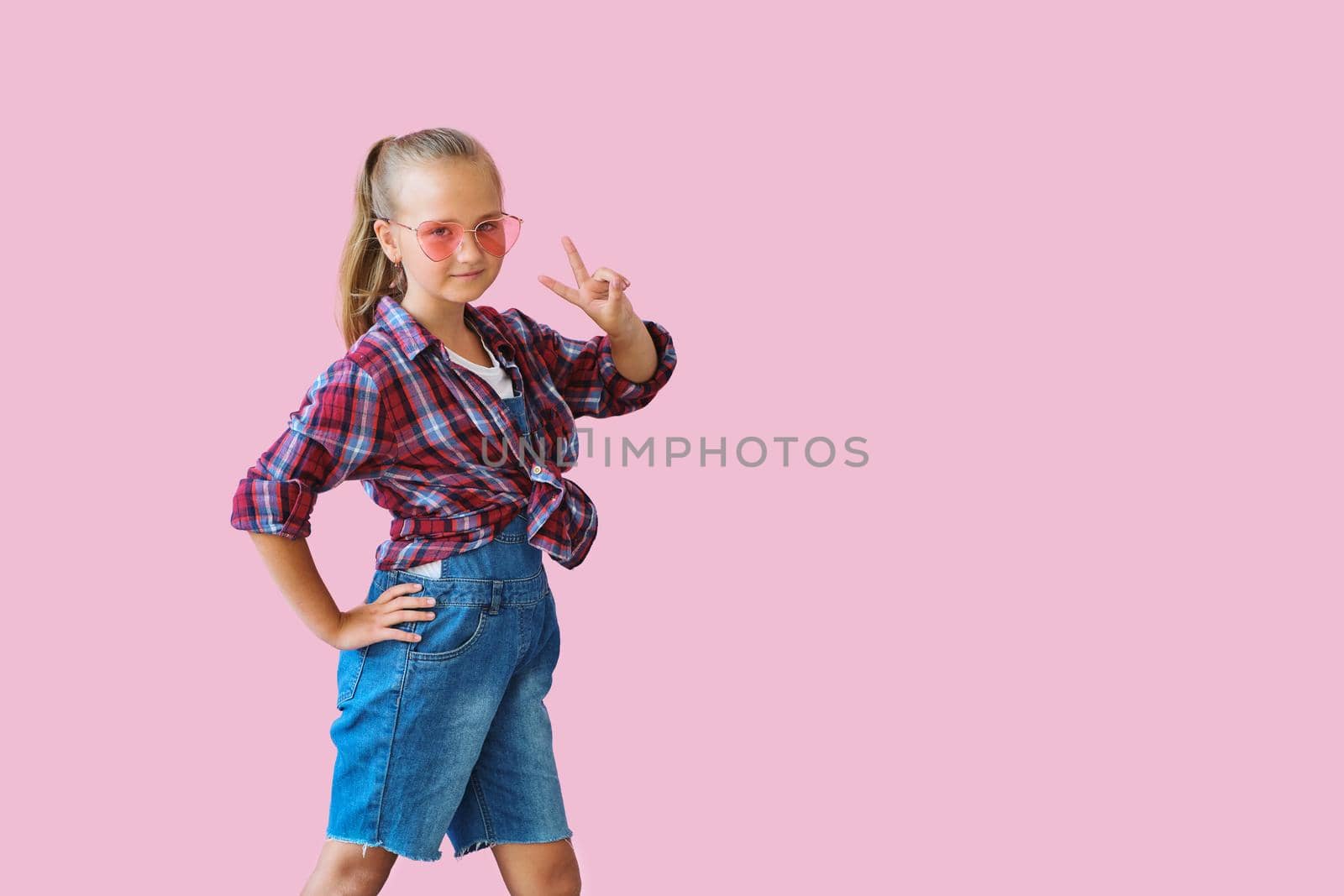 Pretty cool kid girl in pink sunglasses posing against pink background. Fashion style portrait of young happy smiling woman, copy space.