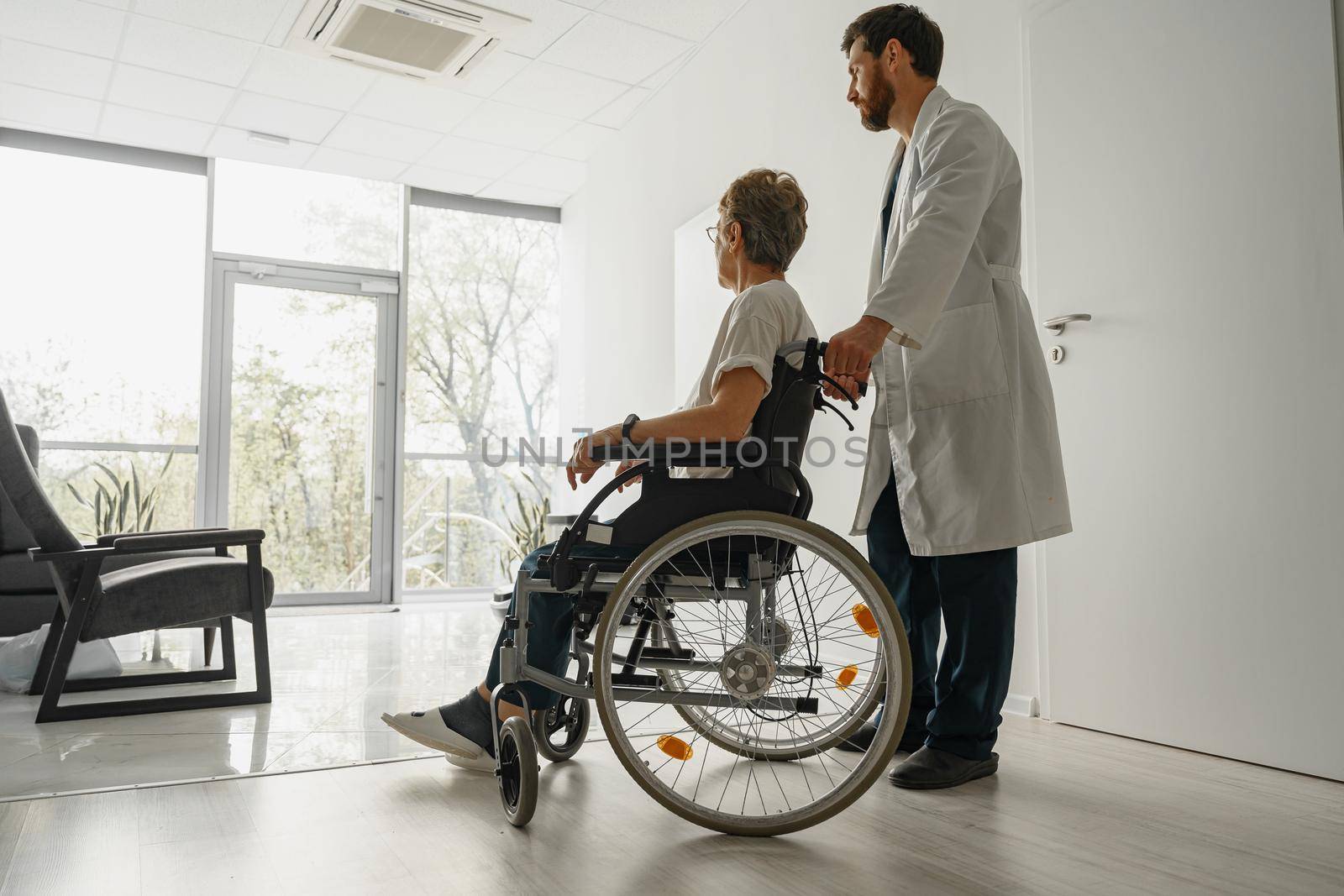 Professional male doctor carrying patient on wheelchair in medicine clinic hall. High quality photo