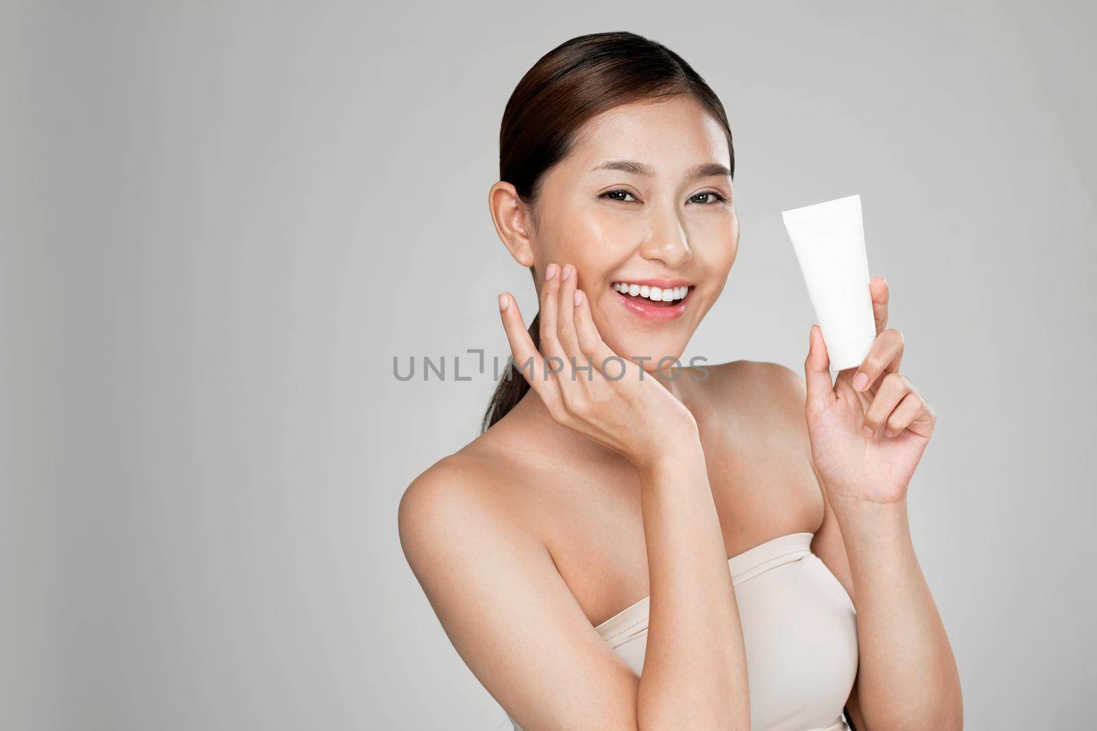 Ardent woman smiling holding mockup product for advertising text place, light grey background. Concept of healthcare for skin, beauty care product for advertising.