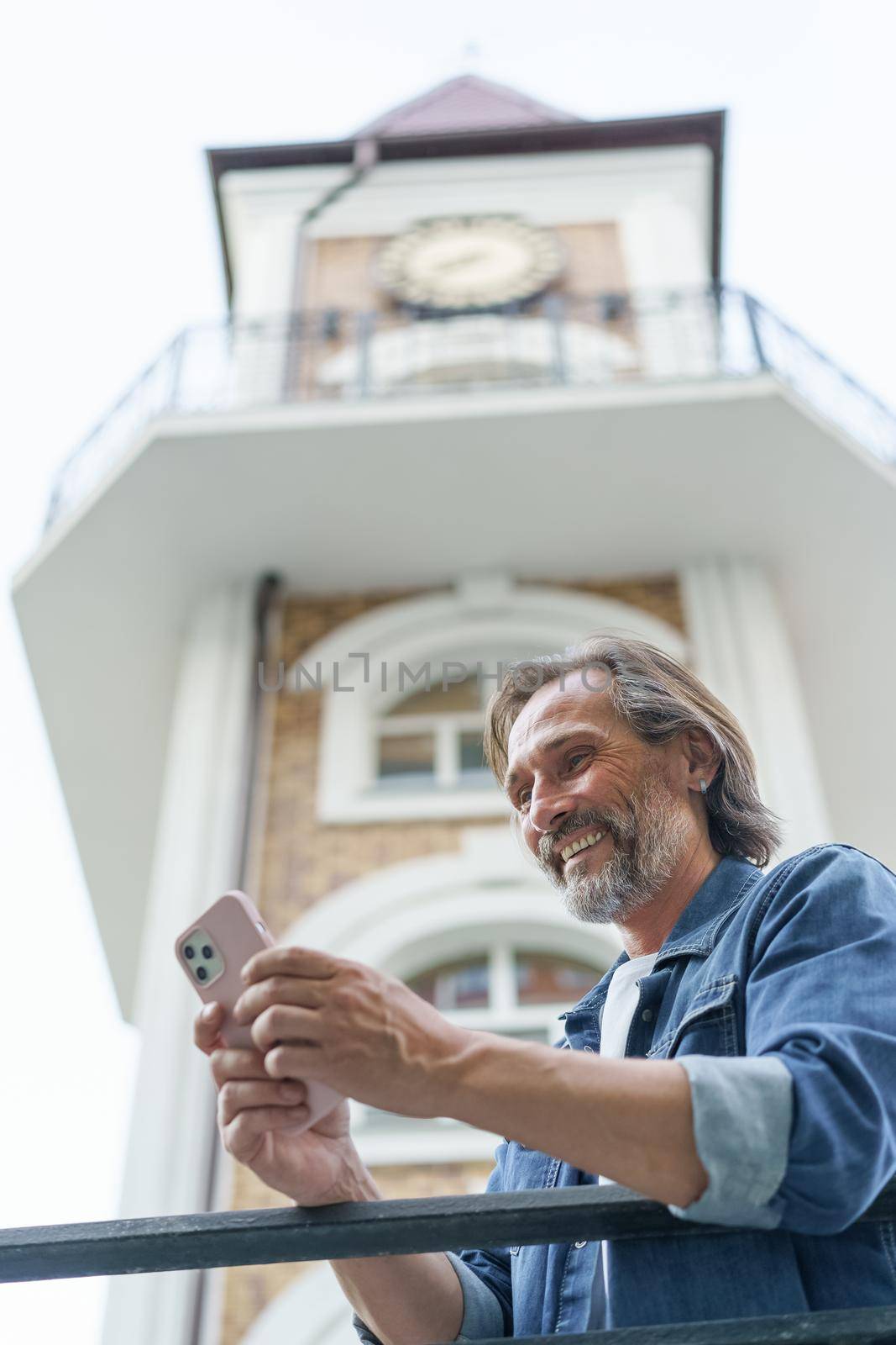 Mature happy grey bearded man laughing reading text message or funny social media looking at smartphone standing in old town wearing casual. Business and travel concept.