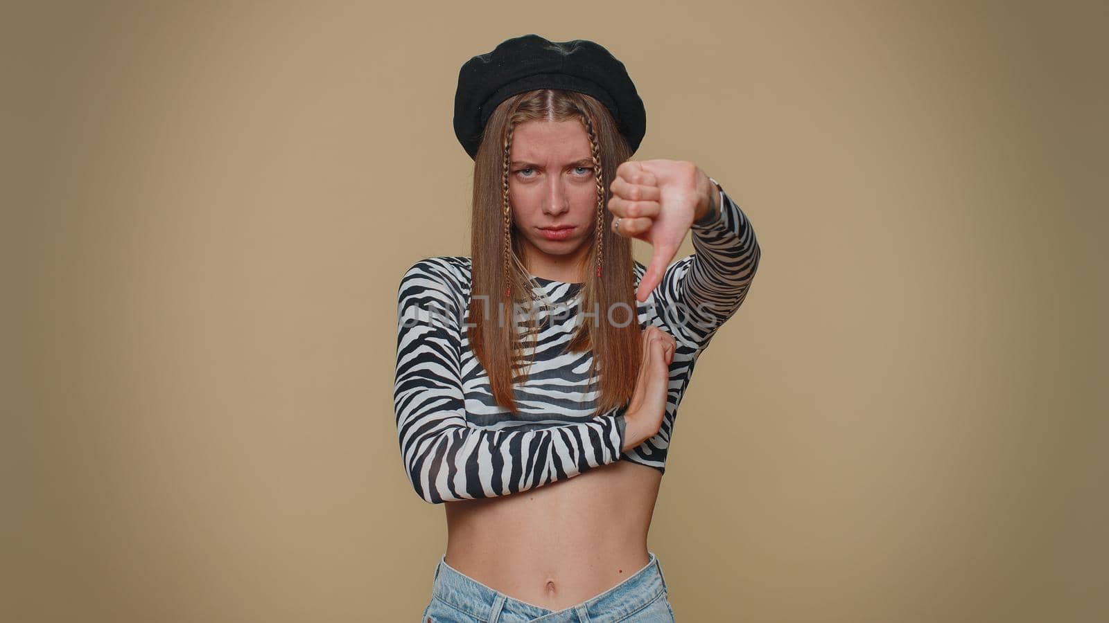 Upset lovely pretty young woman showing thumbs down sign gesture, expressing discontent, disapproval, dissatisfied, dislike. Adult stylish female girl isolated alone on beige studio background indoors