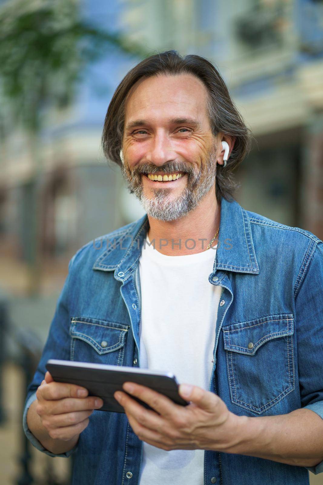 Confident freelancer man with grey haired hold digital tablet in hands having a call while standing on urban streets. Mature man talking or listening music use wireless earphones travel old town.