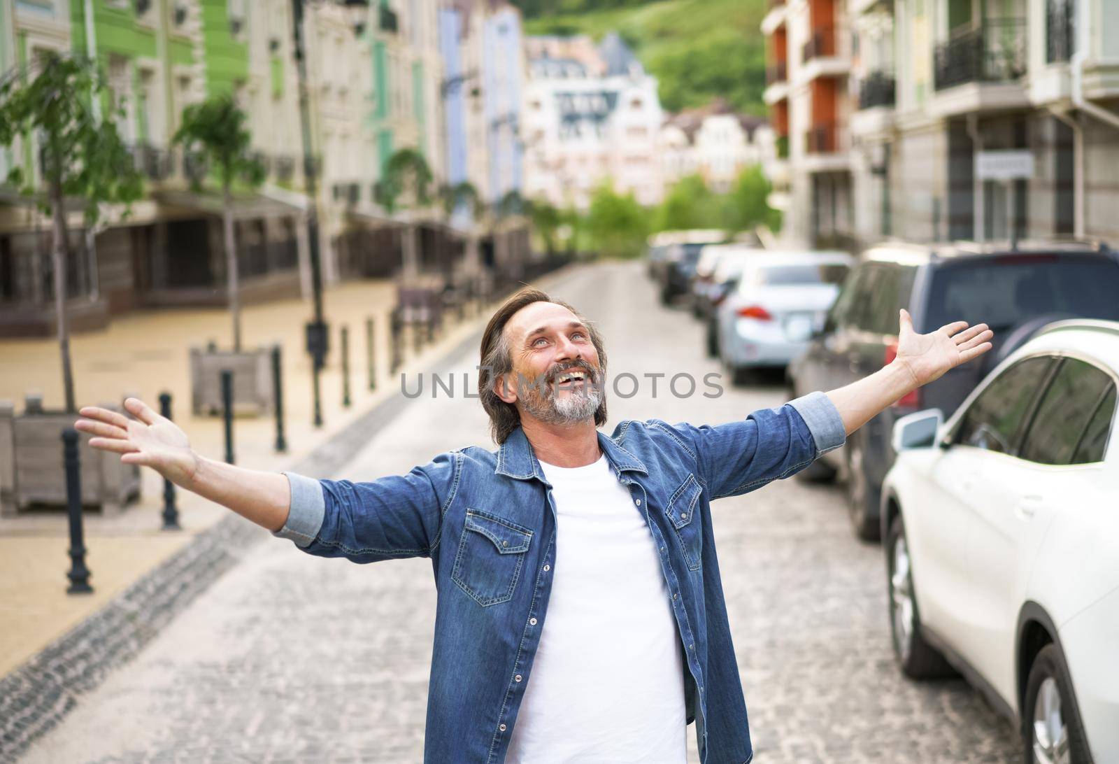 Giving thanks to God mature man standing outdoors with hands spread wide open wearing jeans shirt with white t-shirt under. Mid Aged man glad that life is beautiful standing in old town street by LipikStockMedia