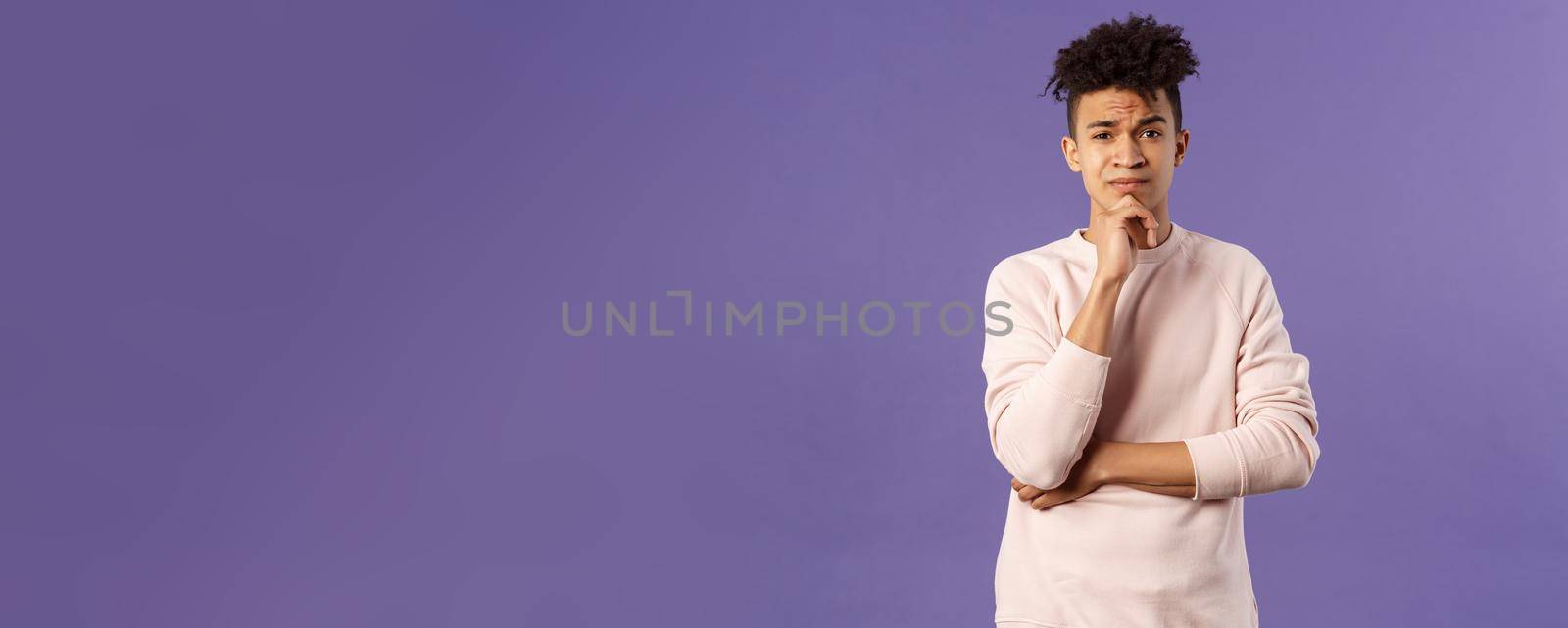 Thoughtful, skeptical young man look judgemental and hesitant at camera while pondering between choices, consider what to do, thinking over purple background, solving troublesome situation.