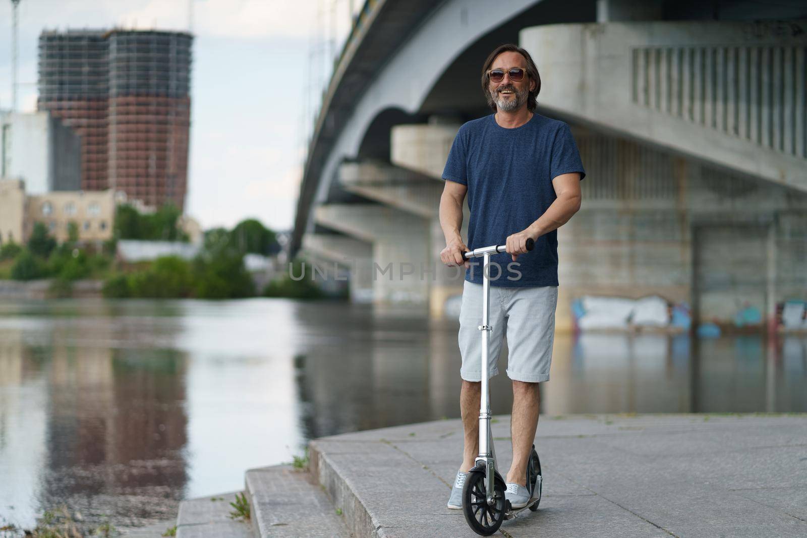 Business on the go. Riding scooter handsome stylish middle aged man with grey beard stand under town bridge over river with urban city on background after work outdoors. Travel, lifestyle concept.