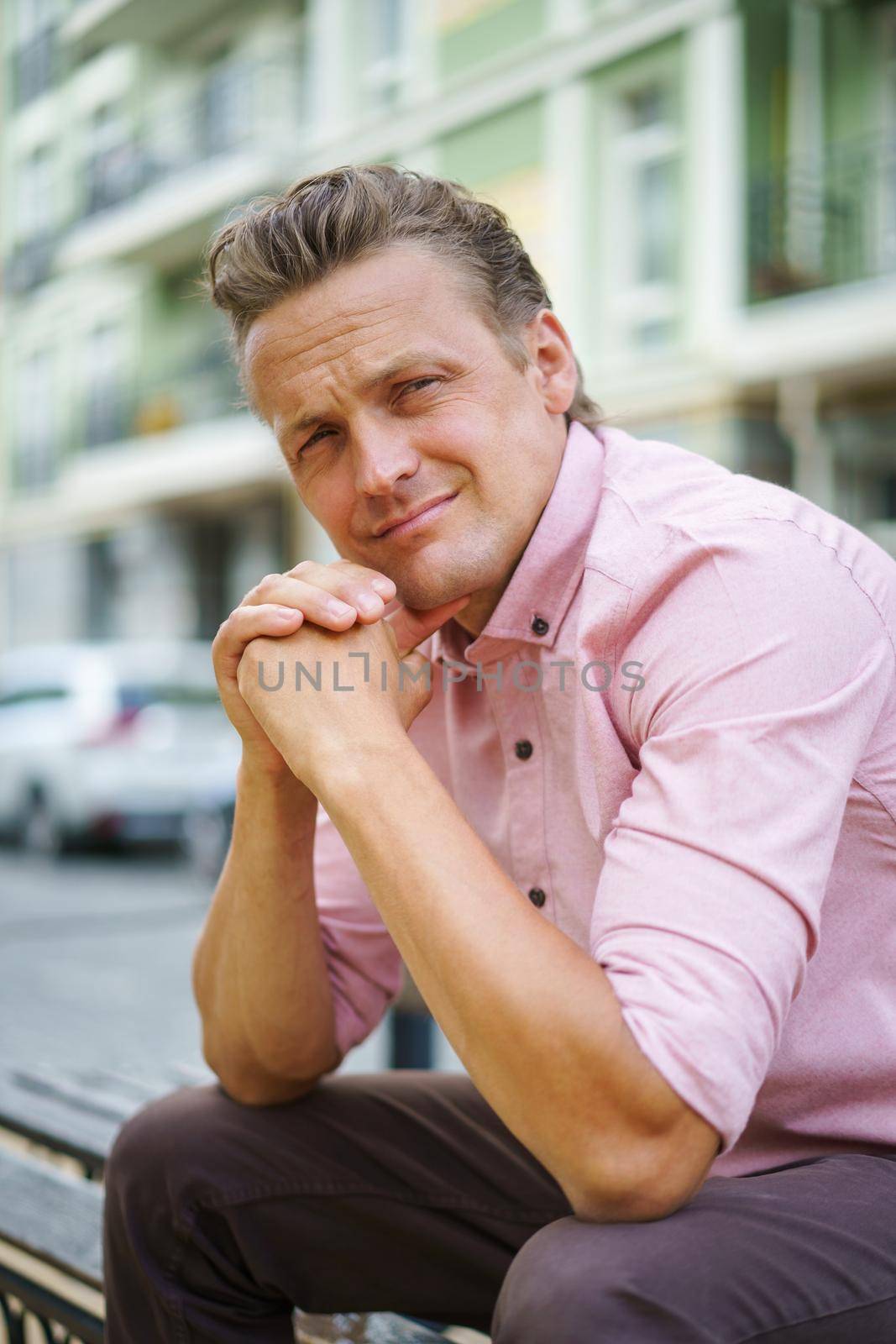 Handsome man spend time sitting on the bench in urban city with hands put together and leaned chin looking at camera, wearing pink shirt. Smiling freelancer caucasian man off work by LipikStockMedia
