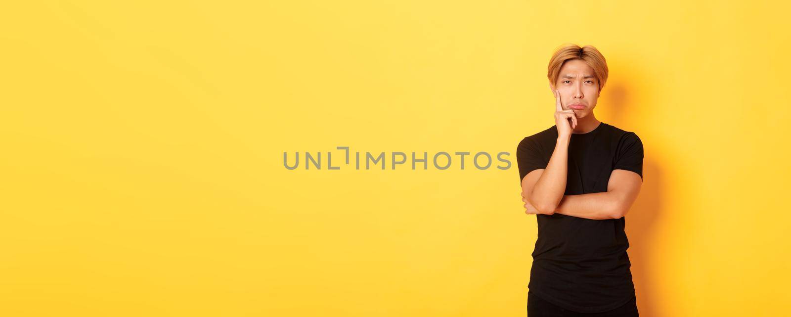 Upset thoughtful asian guy sulking and looking disappointed after thinking, standing yellow background by Benzoix