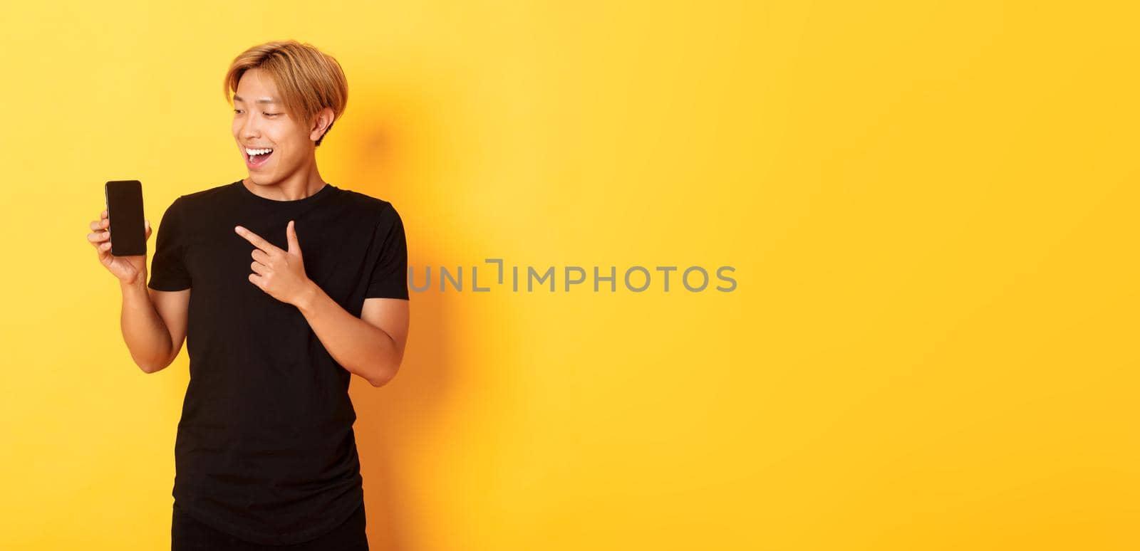 Portrait of satisfied asian guy pointing finger and looking at smartphone screen with pleased smile, showing application, standing yellow background by Benzoix