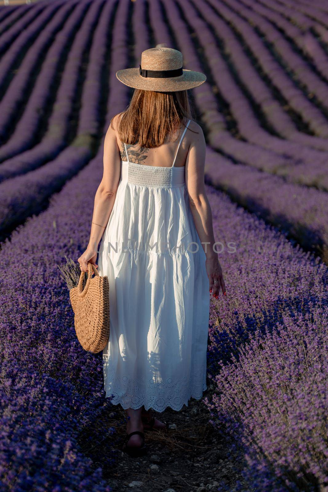 A middle-aged woman, blonde in a white dress and hat, walks through a lavender field with a basket.