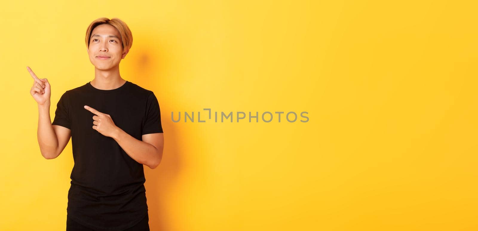 Thoughtful and pleased handsome asian man smiling and pointing fingers upper left corner, standing over yellow background by Benzoix