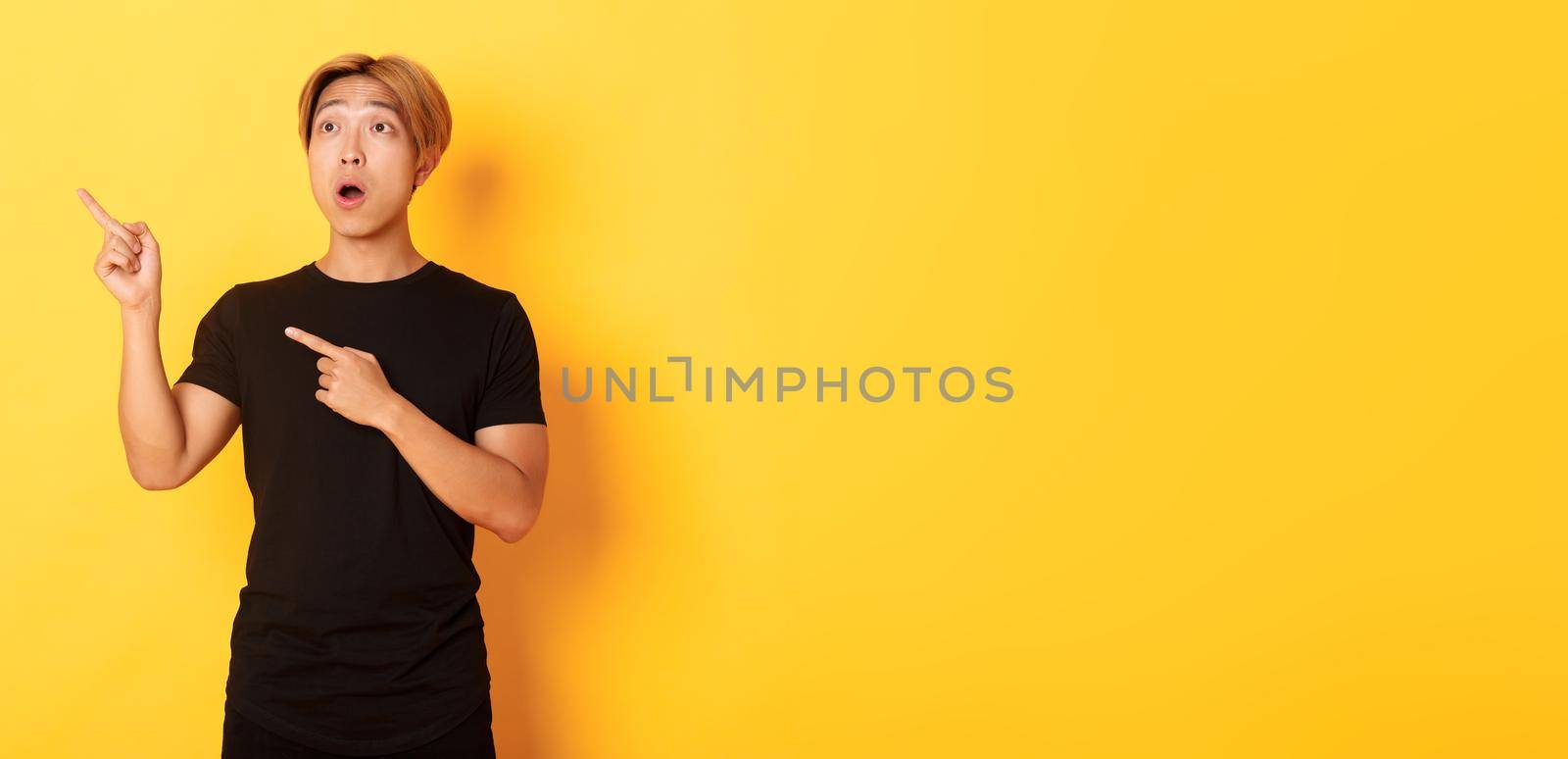 Portrait of impressed asian guy looking and pointing fingers upper left corner, standing yellow background.