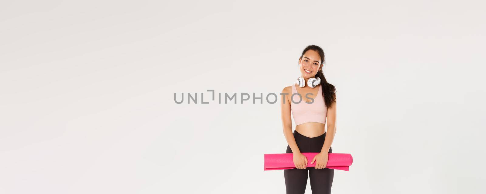 Full length of cute asian girl like fitness, holding rubber mat for exercises or yoga classes, standing in sportswear looking happy after productive training in gym, good workout, white background.
