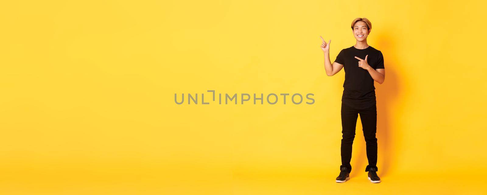 Full-length of smiling attractive korean guy pointing fingers left, showing logo, yellow background.