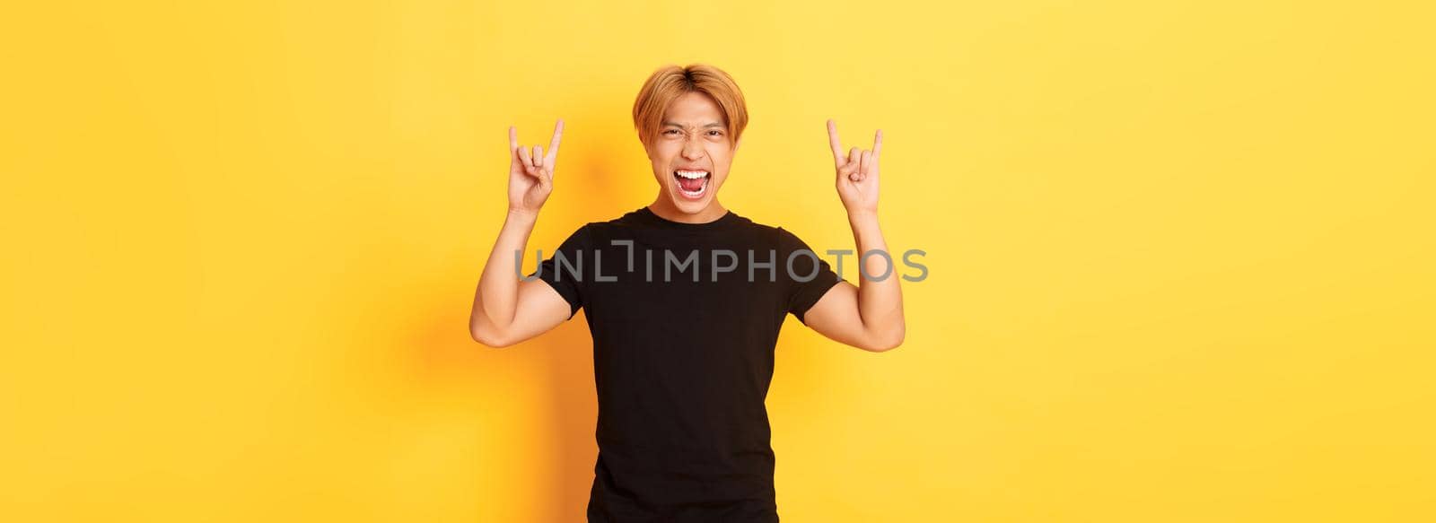Portrait of happy handsome asian man having fun and partying, showing rock-n-roll gesture, standing yellow background by Benzoix